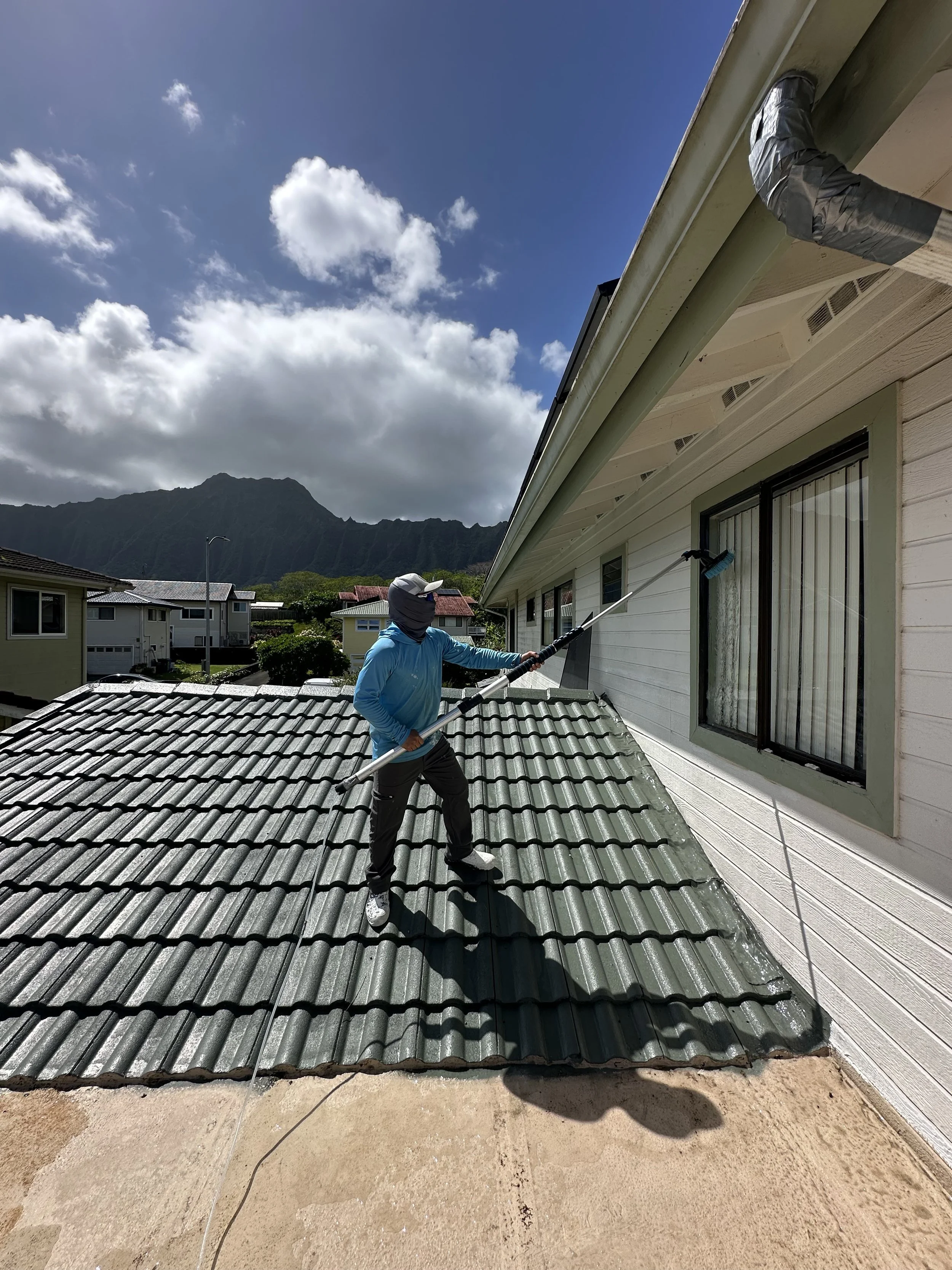 A person cleaning or painting a metal roof on a residential building using a long-handled tool. The house has white siding and a window with curtains. The sky is partly cloudy with a mountain in the background.