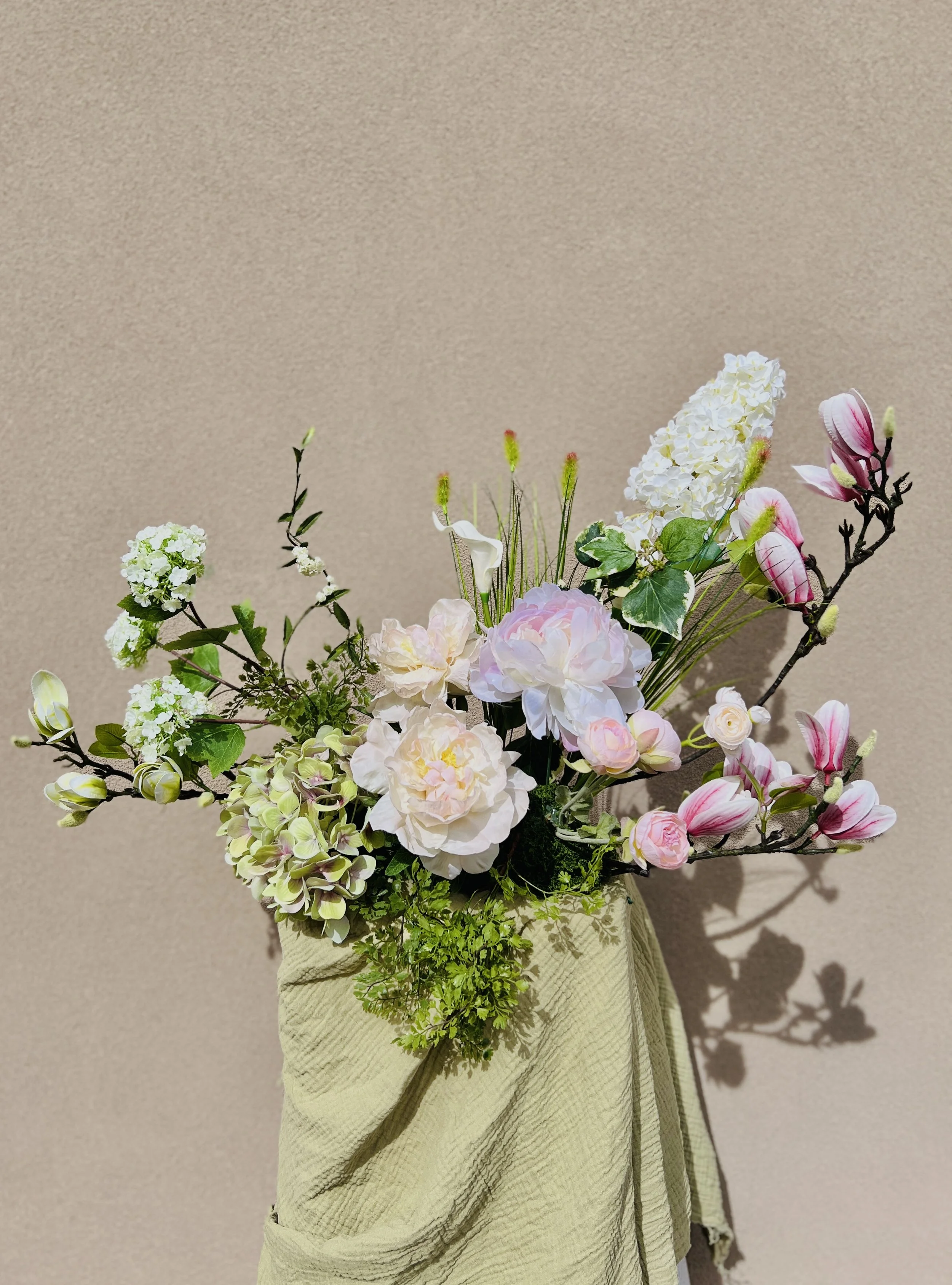 A custom floral arrangement with peonies and magnolias on a green linen draped pedestal against a beige wall.