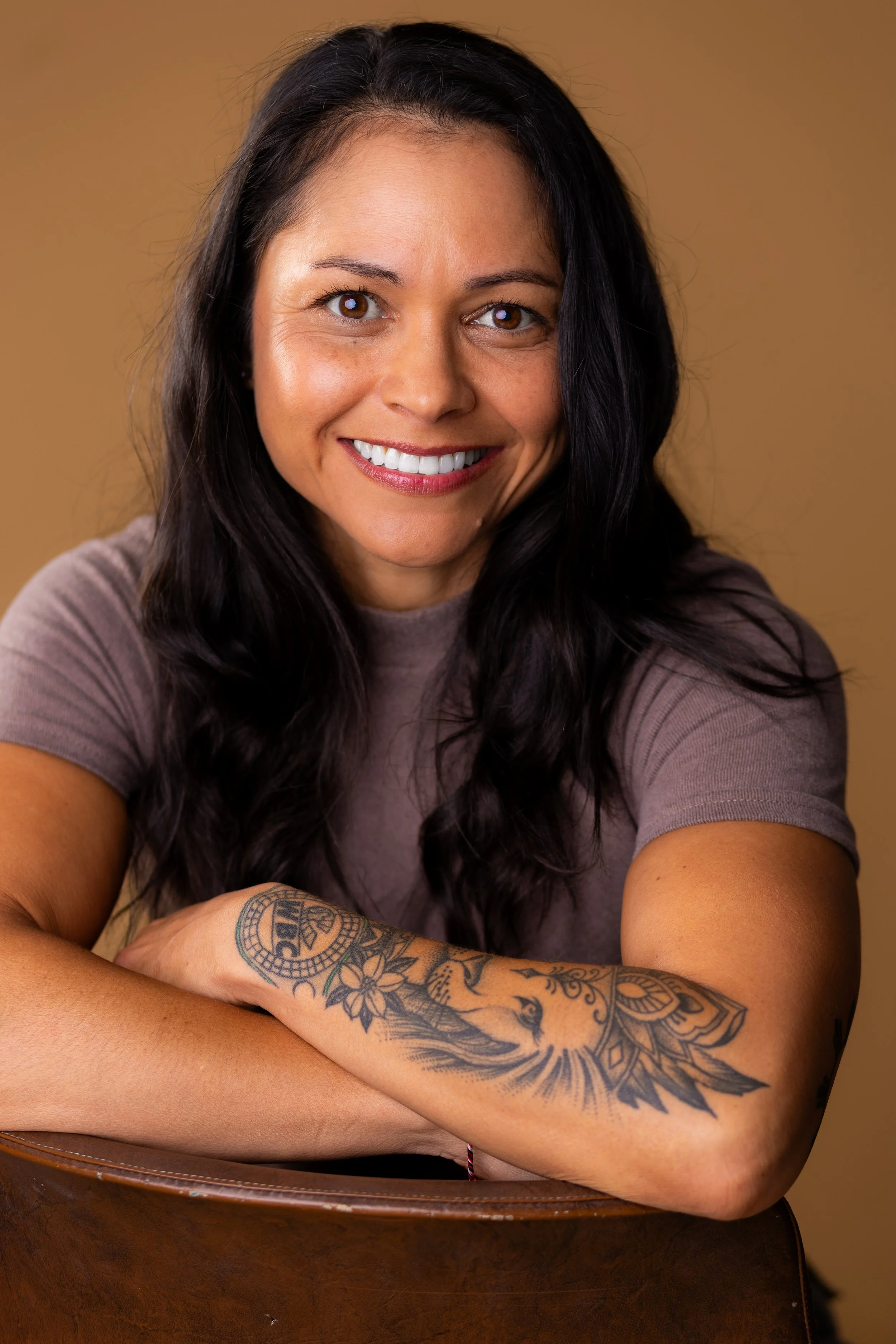 A woman with dark hair and a tattoo on her arm, smiling at the camera, wearing a light brown shirt, against a plain background.