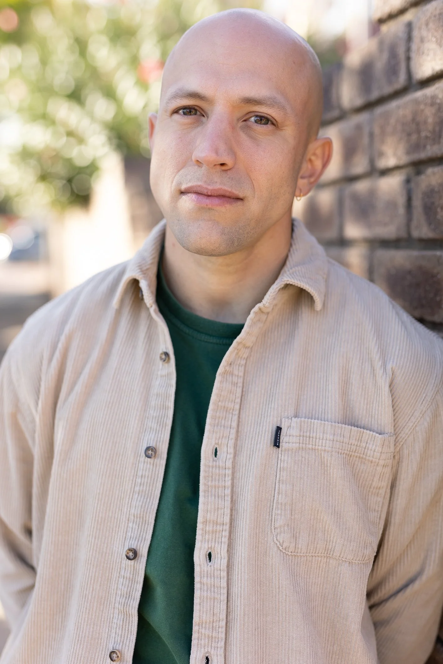 A bald man with light skin and blue eyes wearing a beige corduroy shirt over a dark green t-shirt, standing outdoors against a brick wall with blurred greenery in the background.