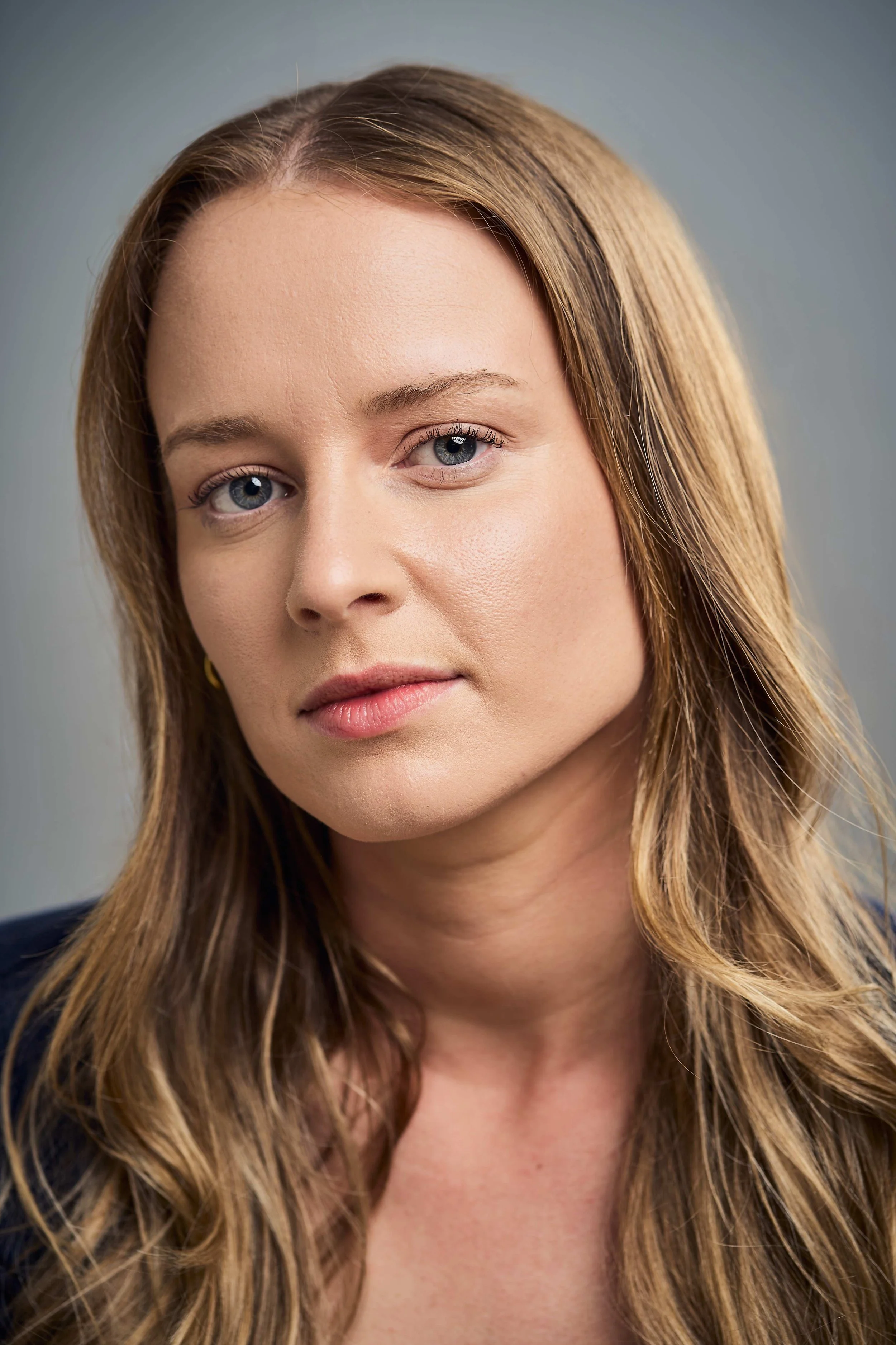 Close-up of a young woman with blonde hair, blue eyes, wearing a navy blue shirt and a small gold necklace, smiling softly at the camera.