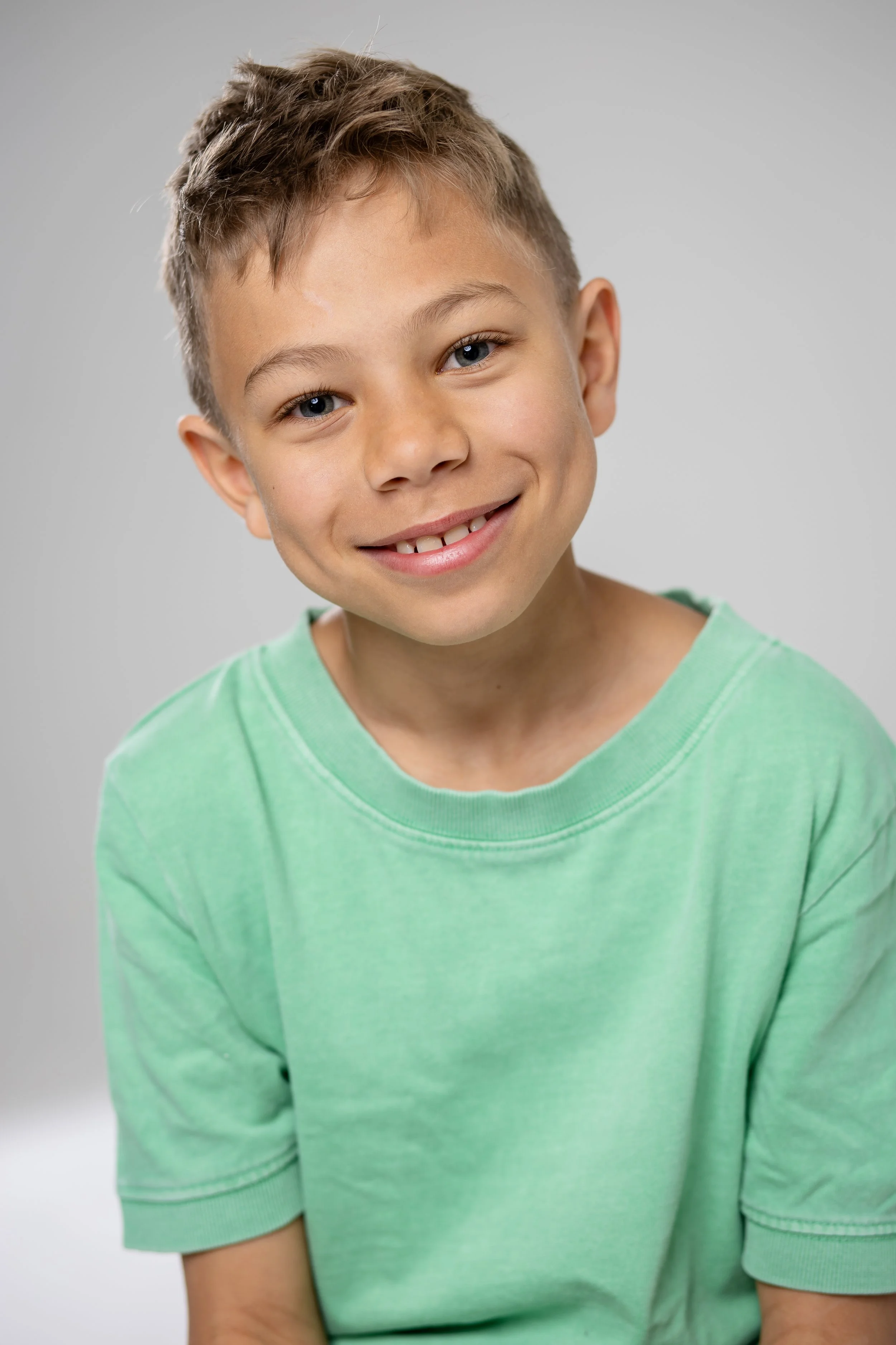 A young boy with light brown hair, blue eyes, and a missing front tooth, smiling at the camera. He's wearing a light green t-shirt.