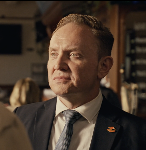 Close-up of a man in a suit and tie, with a serious expression, indoors with blurred background.