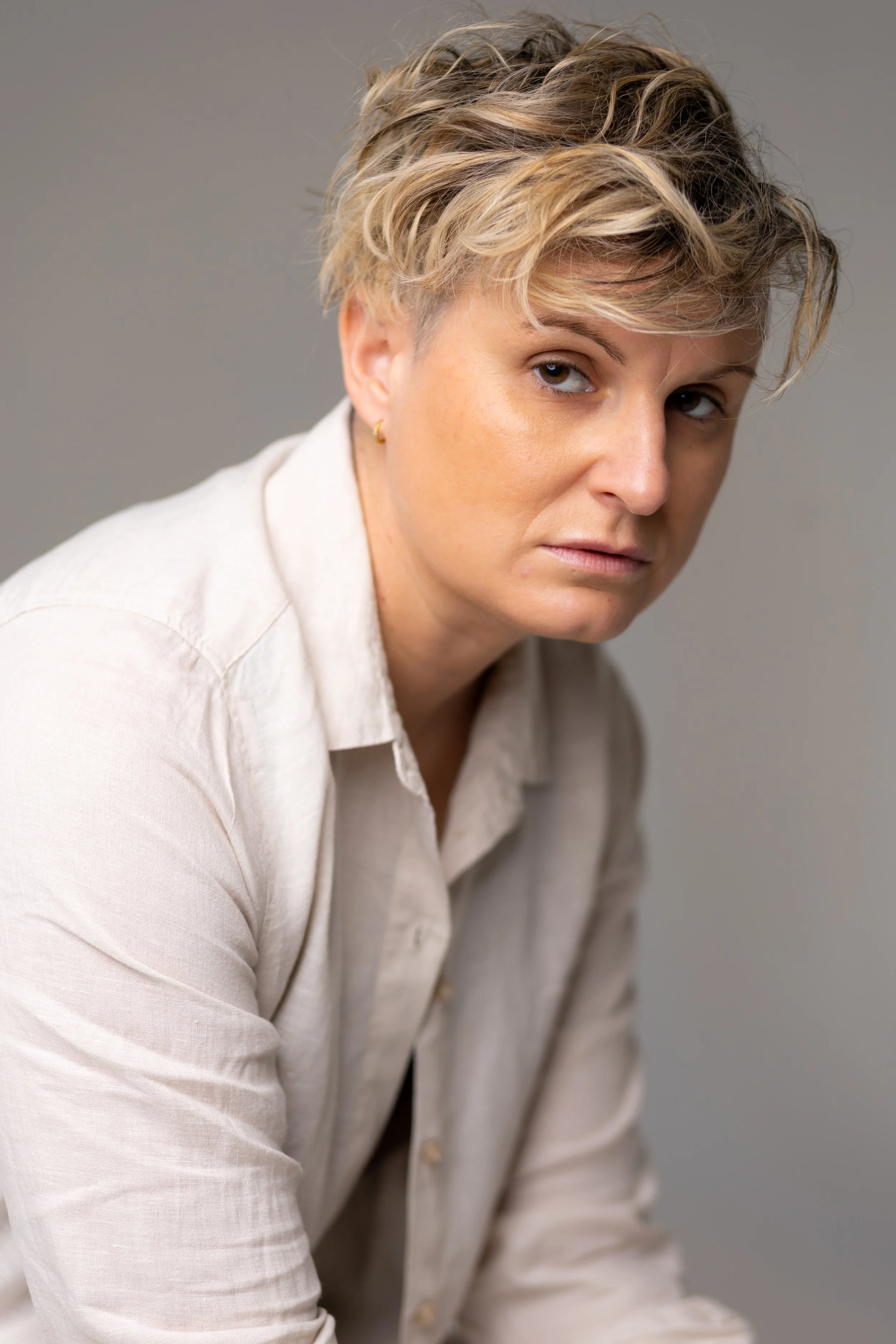 A woman with short, wavy blonde hair looking sideways at the camera, wearing a light-colored shirt, with a neutral gray background.