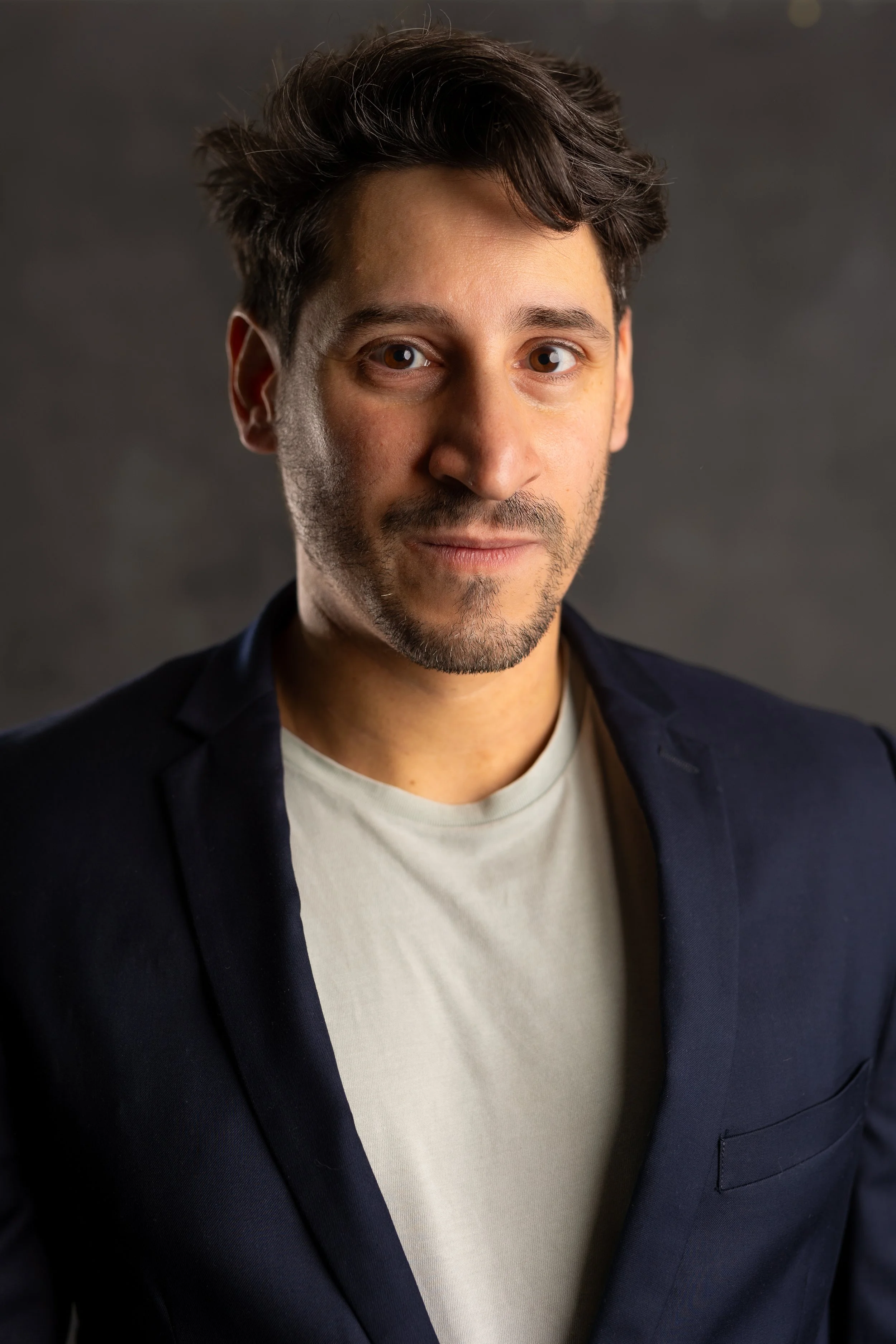 A young man with brown hair and facial hair, wearing a navy blazer and a white t-shirt, looking at the camera with a neutral expression against a dark background.