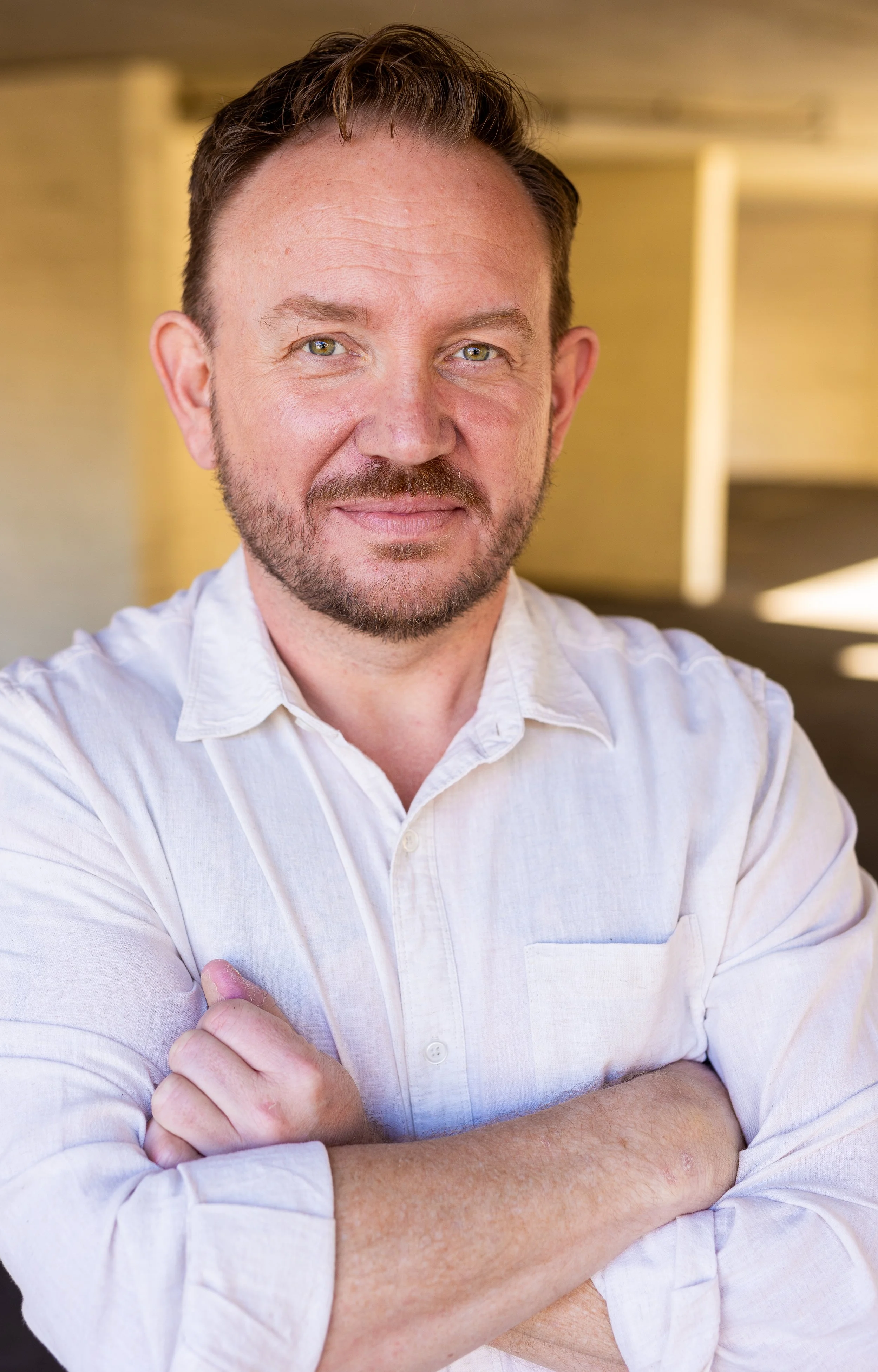 A man with light skin, short brown hair, and a beard wearing a white button-up shirt, standing with arms crossed, smiling in a well-lit indoor setting.