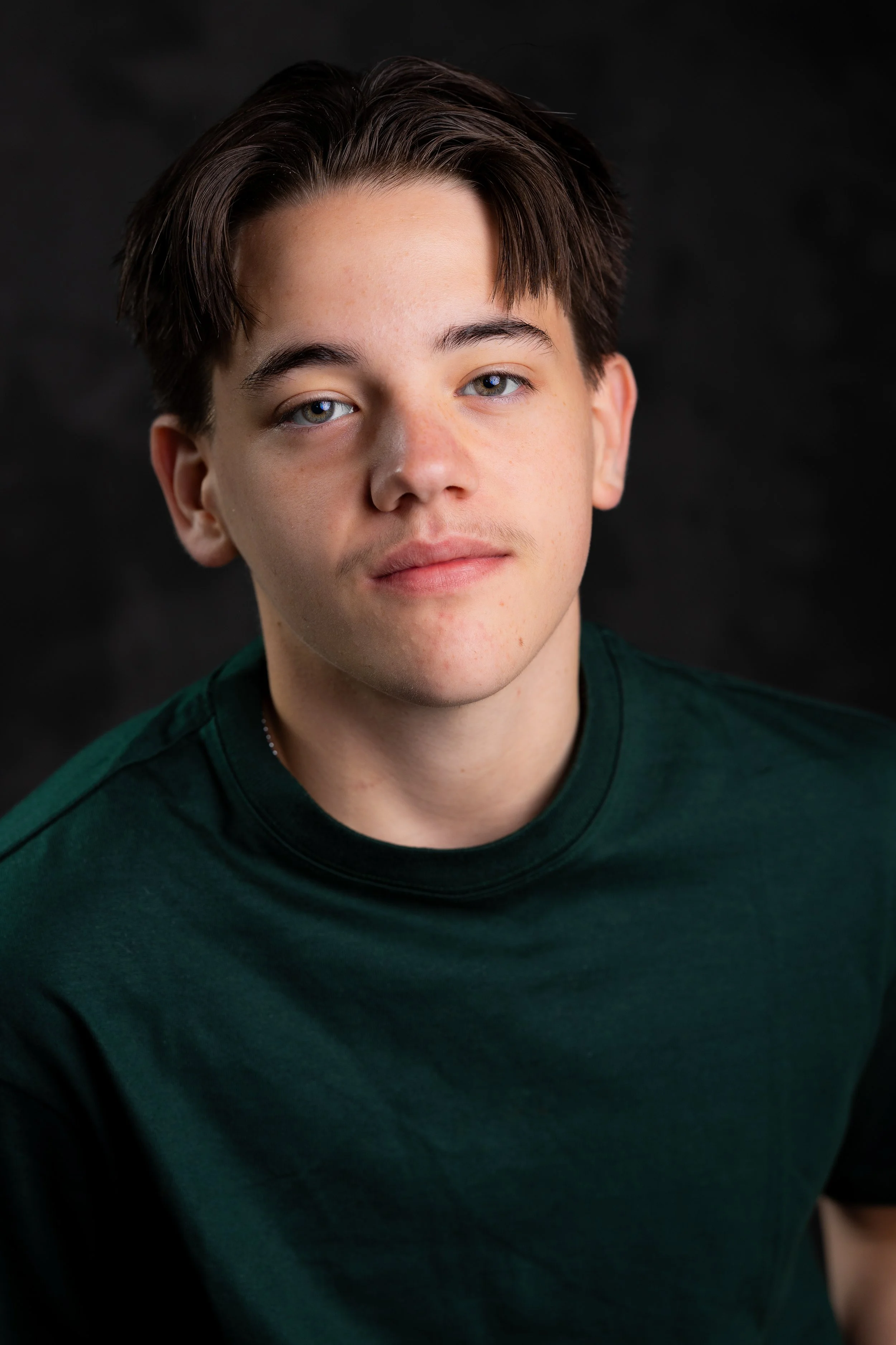 A young man with light skin and brown hair with blue eyes, wearing a dark green shirt, looking at the camera against a black background.