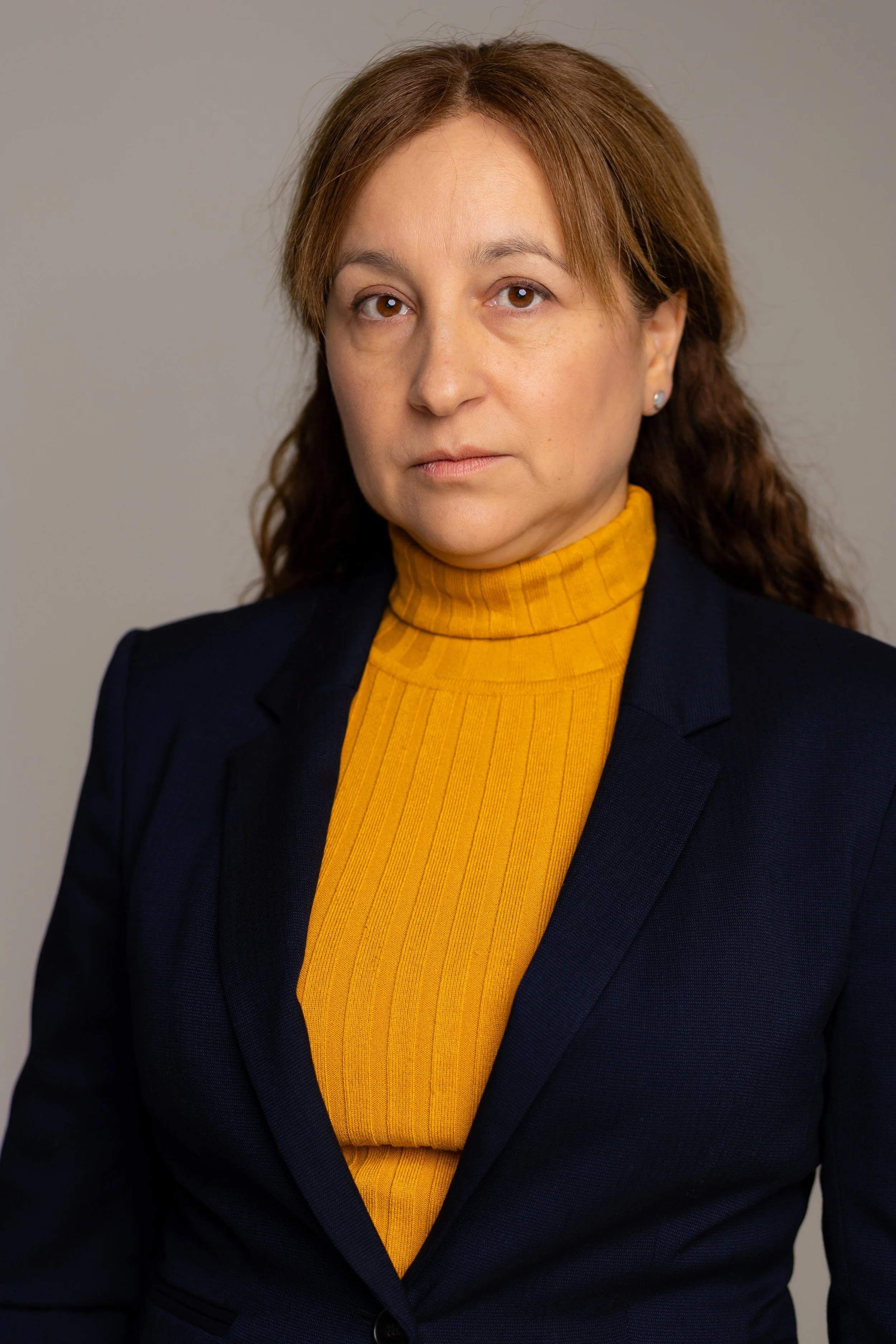 A woman with brown hair wearing a navy blazer and yellow turtleneck, looking directly at the camera.