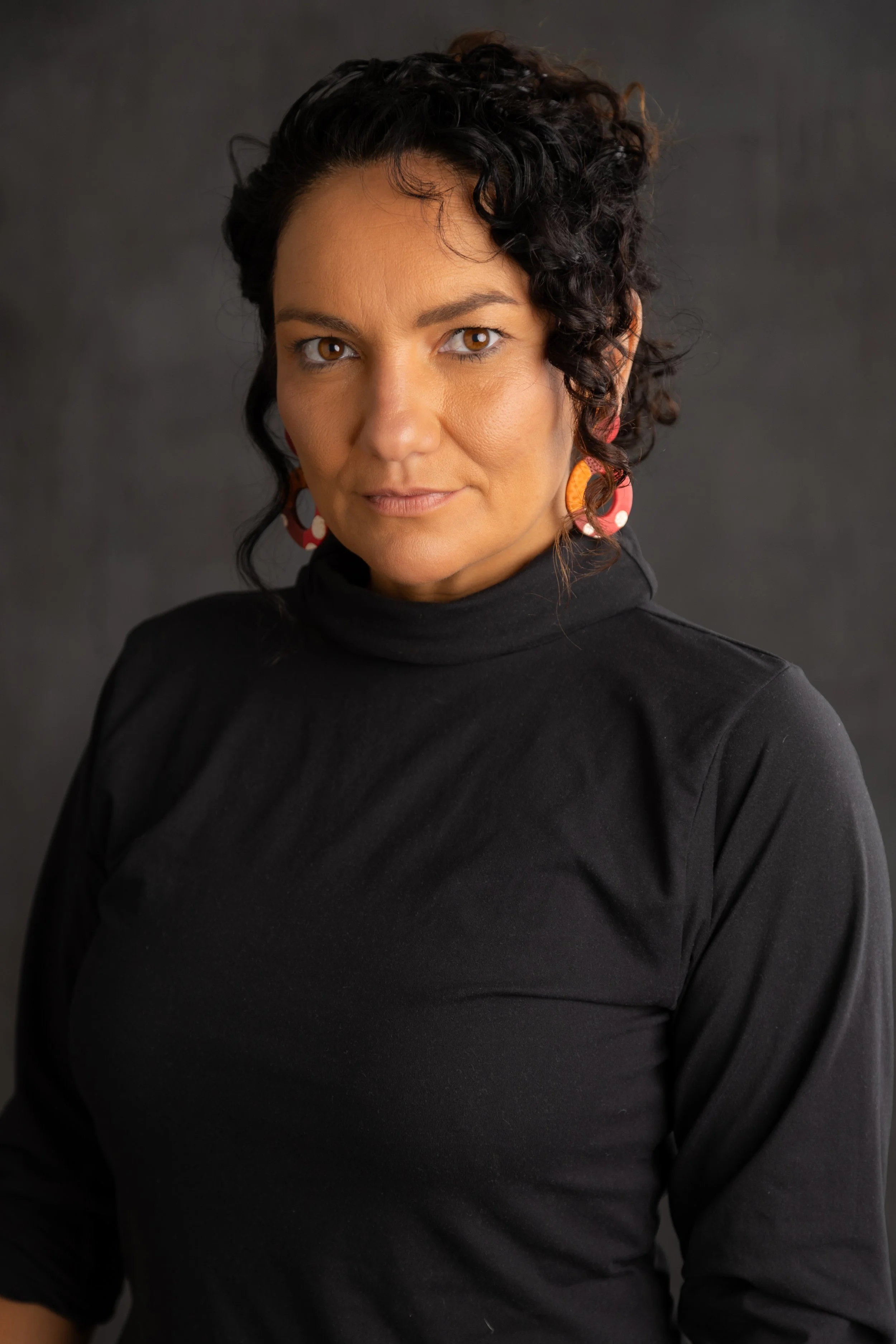 A woman with dark, curly hair wearing a black turtleneck and colorful earrings against a dark background.