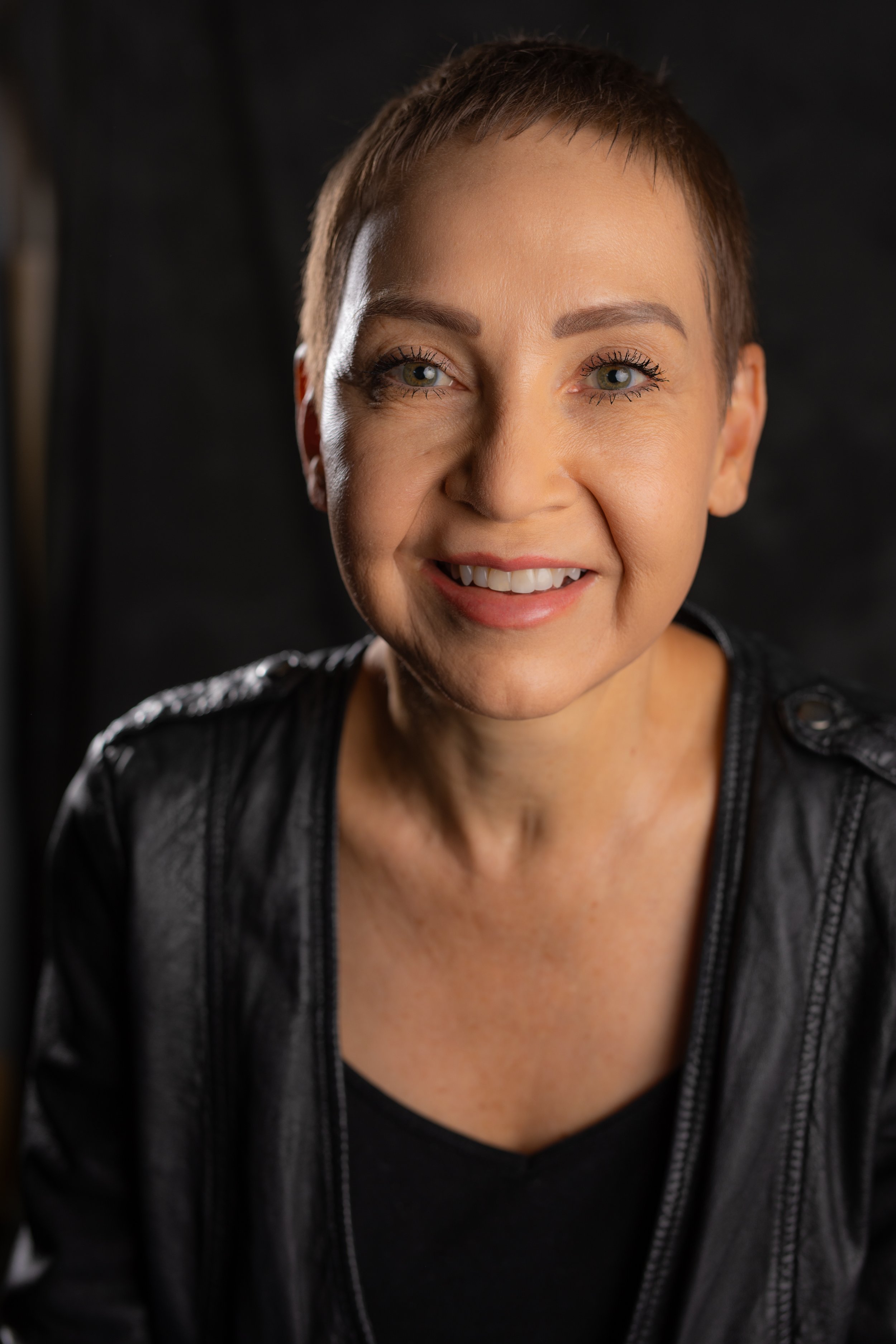 A smiling woman with short brown hair, wearing a black leather jacket, against a dark background.