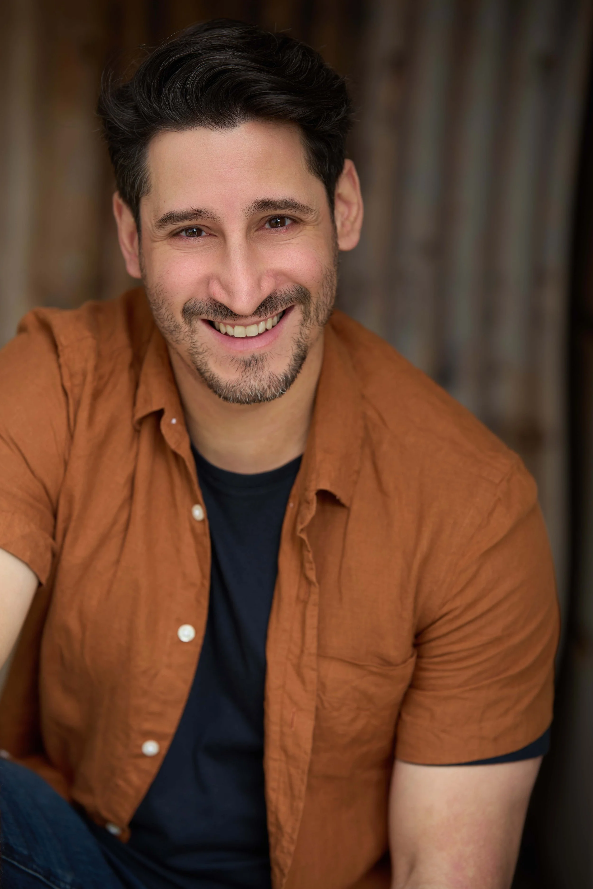 Portrait of a man with brown hair, light facial hair, wearing a white t-shirt and a navy blazer over a dark background.