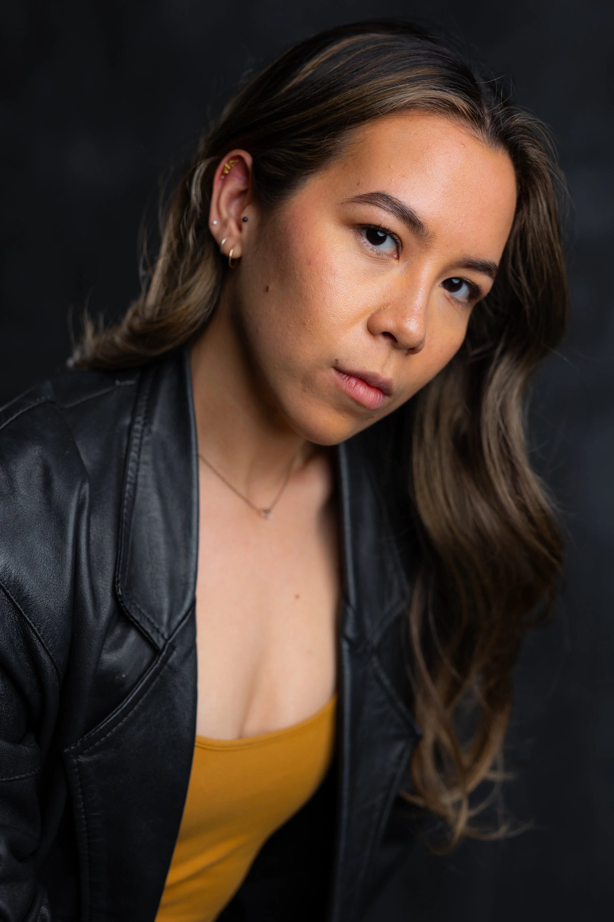A young woman with long wavy brown hair, wearing a black leather jacket over a mustard yellow top, looking directly at the camera against a dark background.