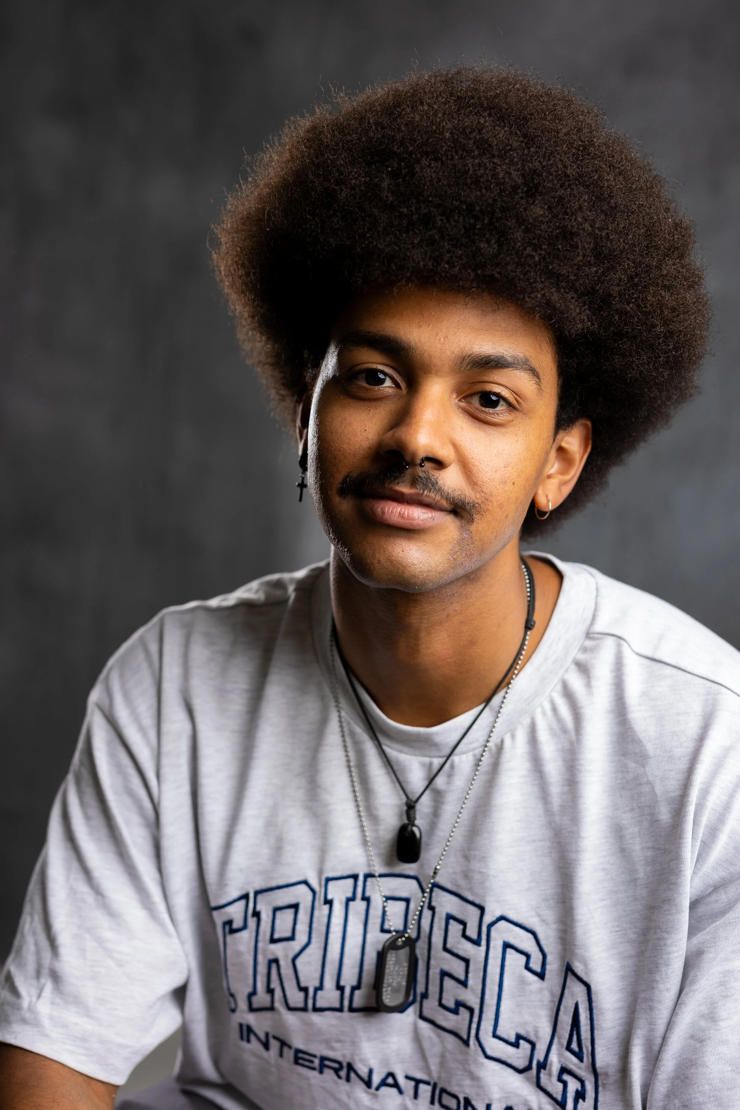 Portrait of a young man with an Afro hairstyle, wearing a white T-shirt with 'TRI-SEC' printed on it, layered necklaces, earrings, and a nose piercing, against a dark blurred background.