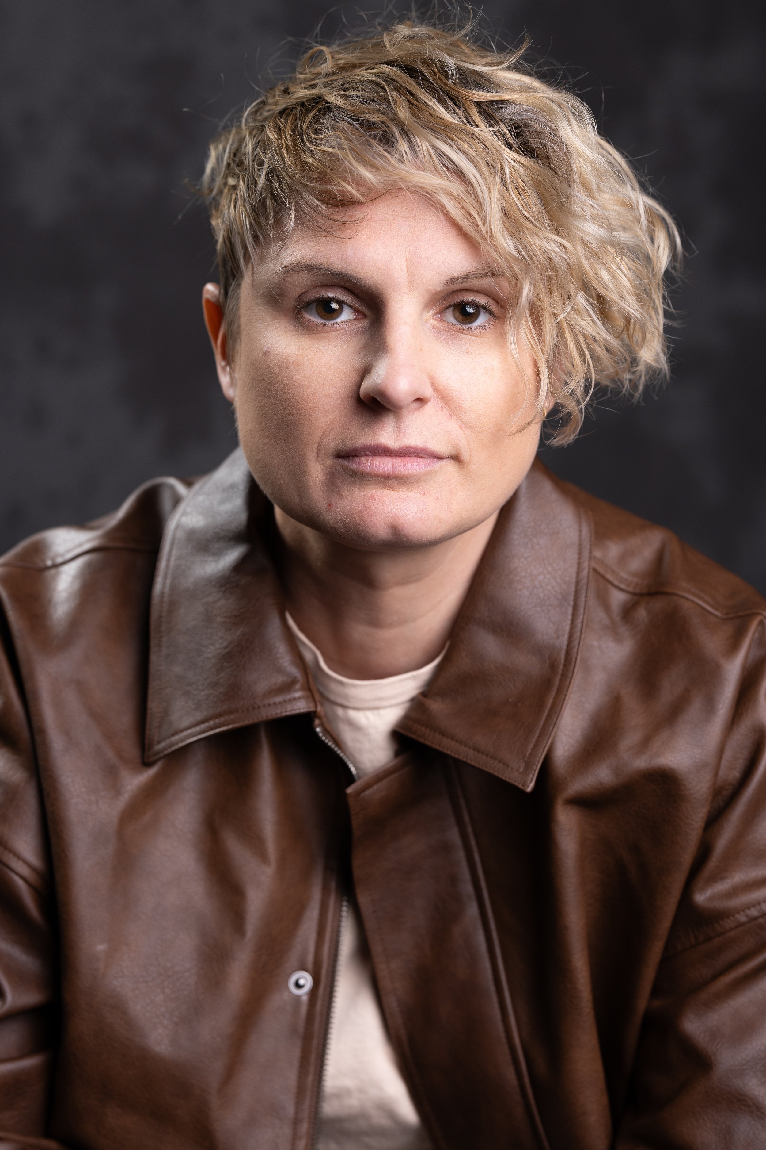 A woman with short, curly blonde hair wearing a brown leather jacket against a dark background.