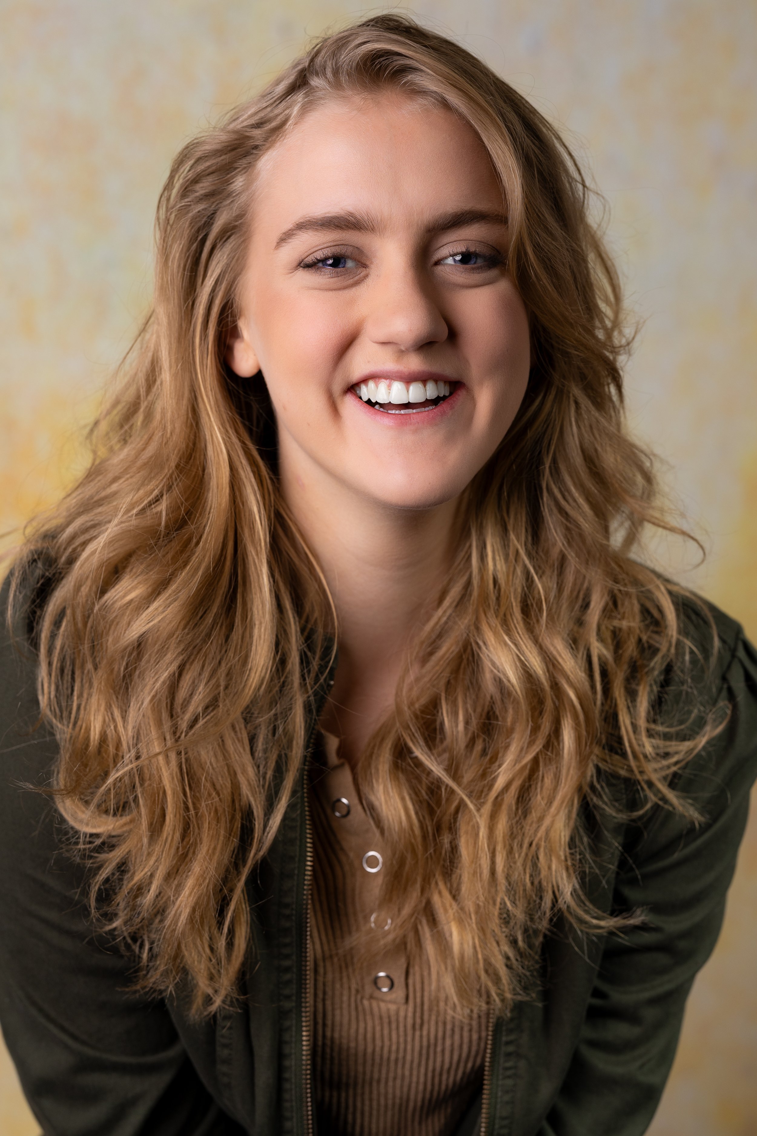 A young woman with long, wavy, light brown hair smiling at the camera, wearing a dark jacket over a brown top with small metallic embellishments, against a beige background.