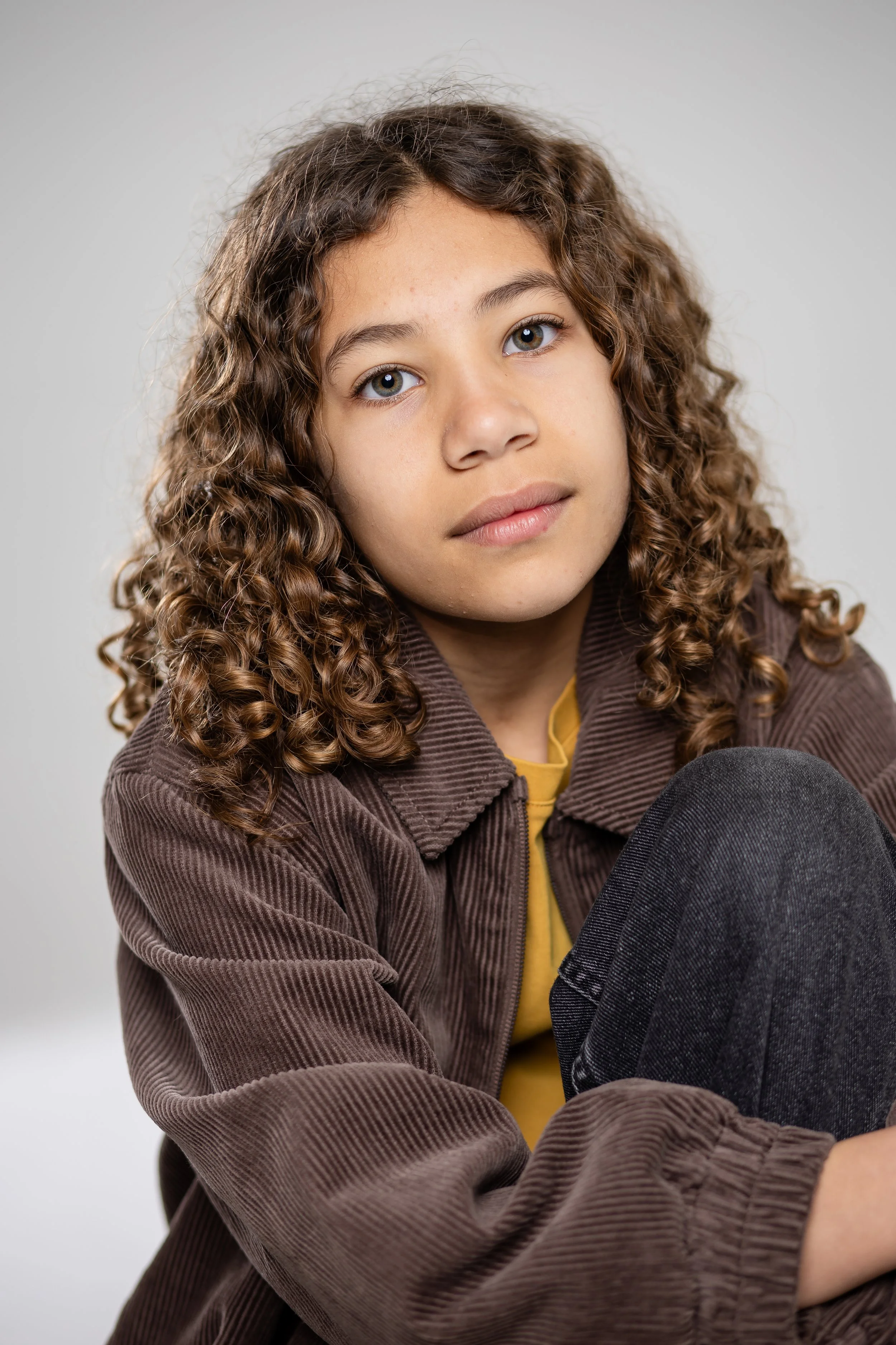 A young girl with curly brown hair and blue eyes wearing a brown corduroy jacket and yellow shirt, sitting on the floor with a neutral expression against a plain light gray background.