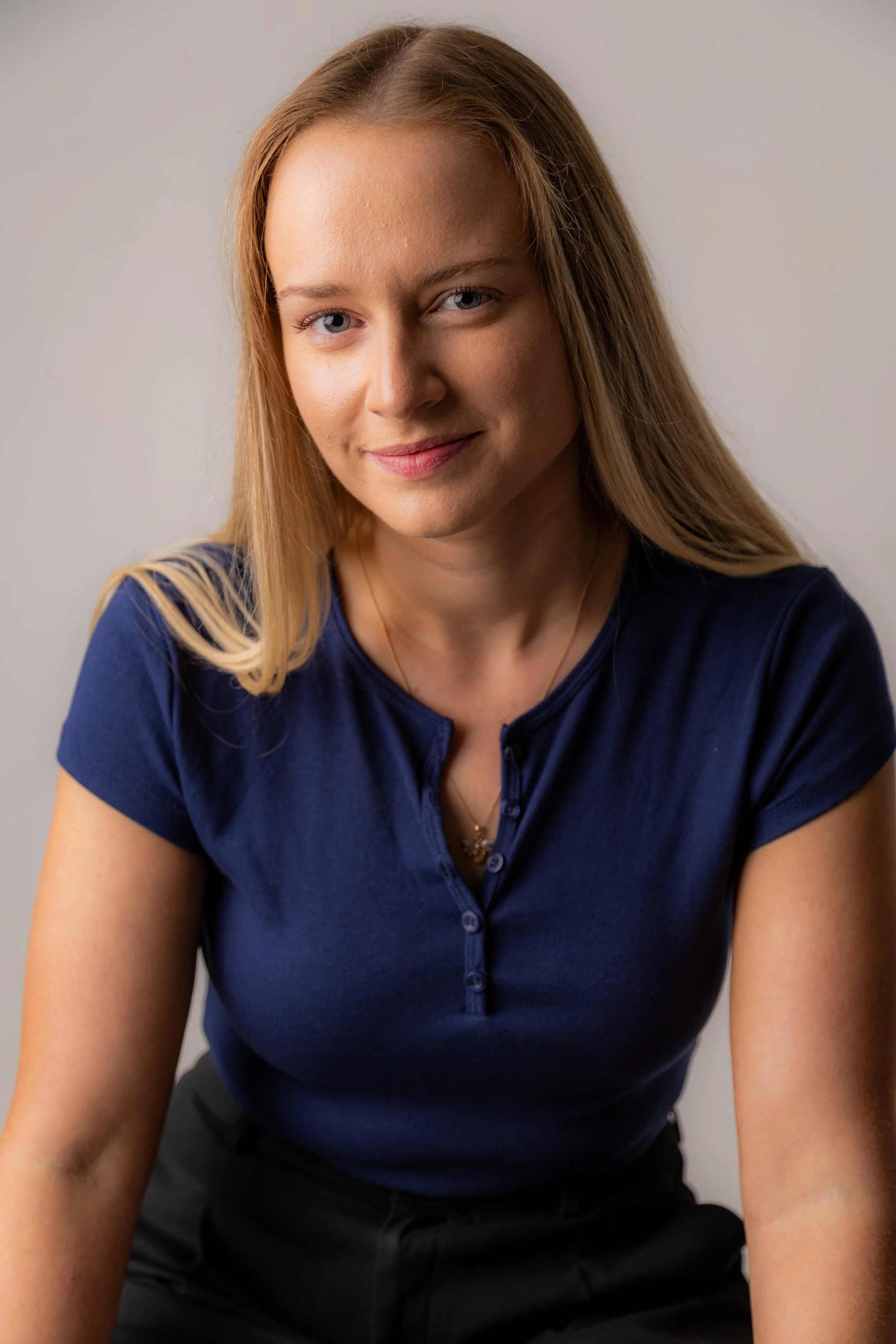 A woman with long blonde hair, wearing a blue short-sleeved shirt, smiling softly at the camera against a plain background.