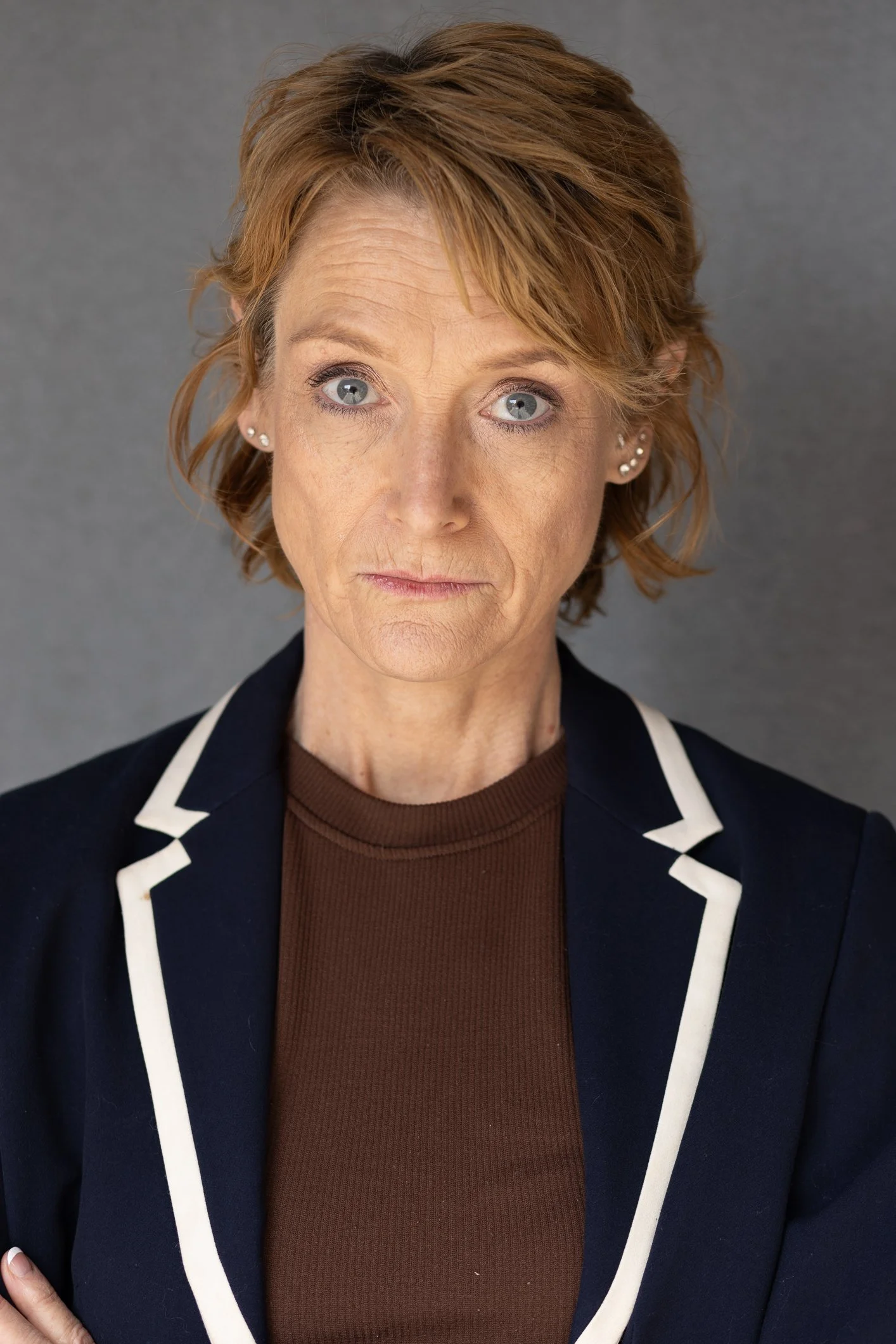 A portrait of a woman with short red hair, blue eyes, and light skin wearing a dark blazer with white trim and a brown top, standing against a gray background.