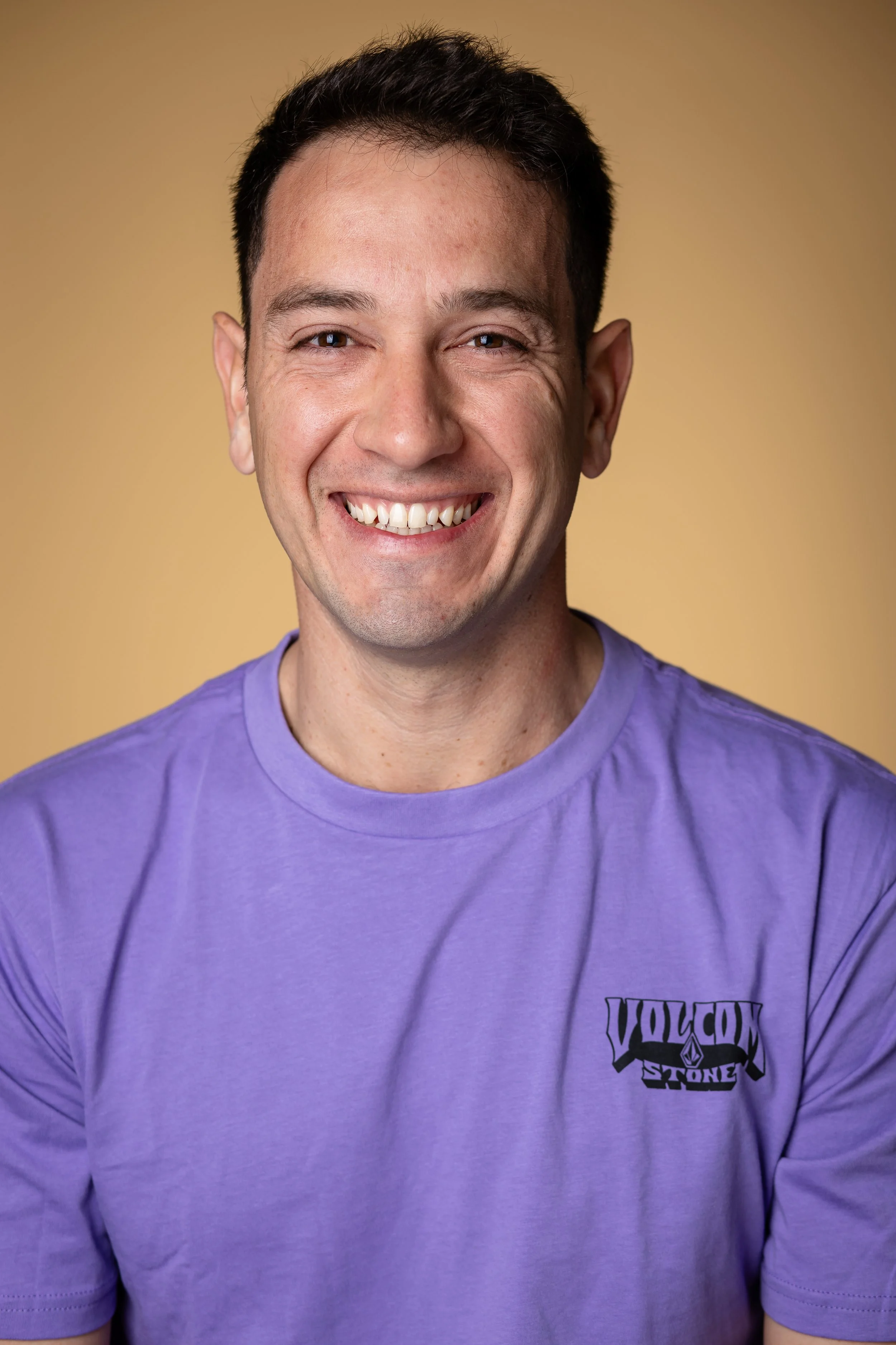 A smiling man with short dark hair wearing a purple T-shirt, standing against a beige background.