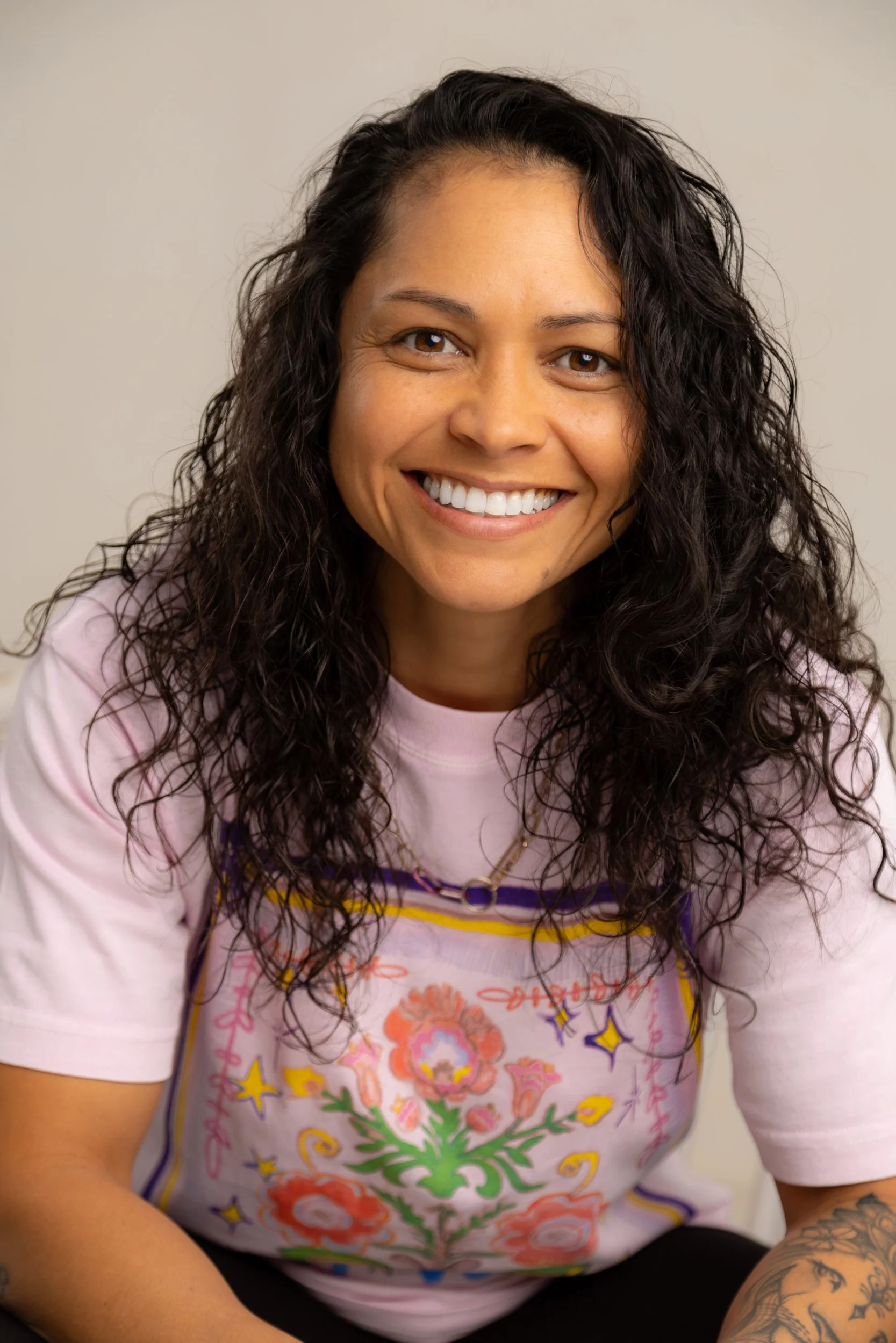 A smiling woman with curly dark hair wearing a pink T-shirt with colorful floral and star designs, and a tattoo on her right arm.