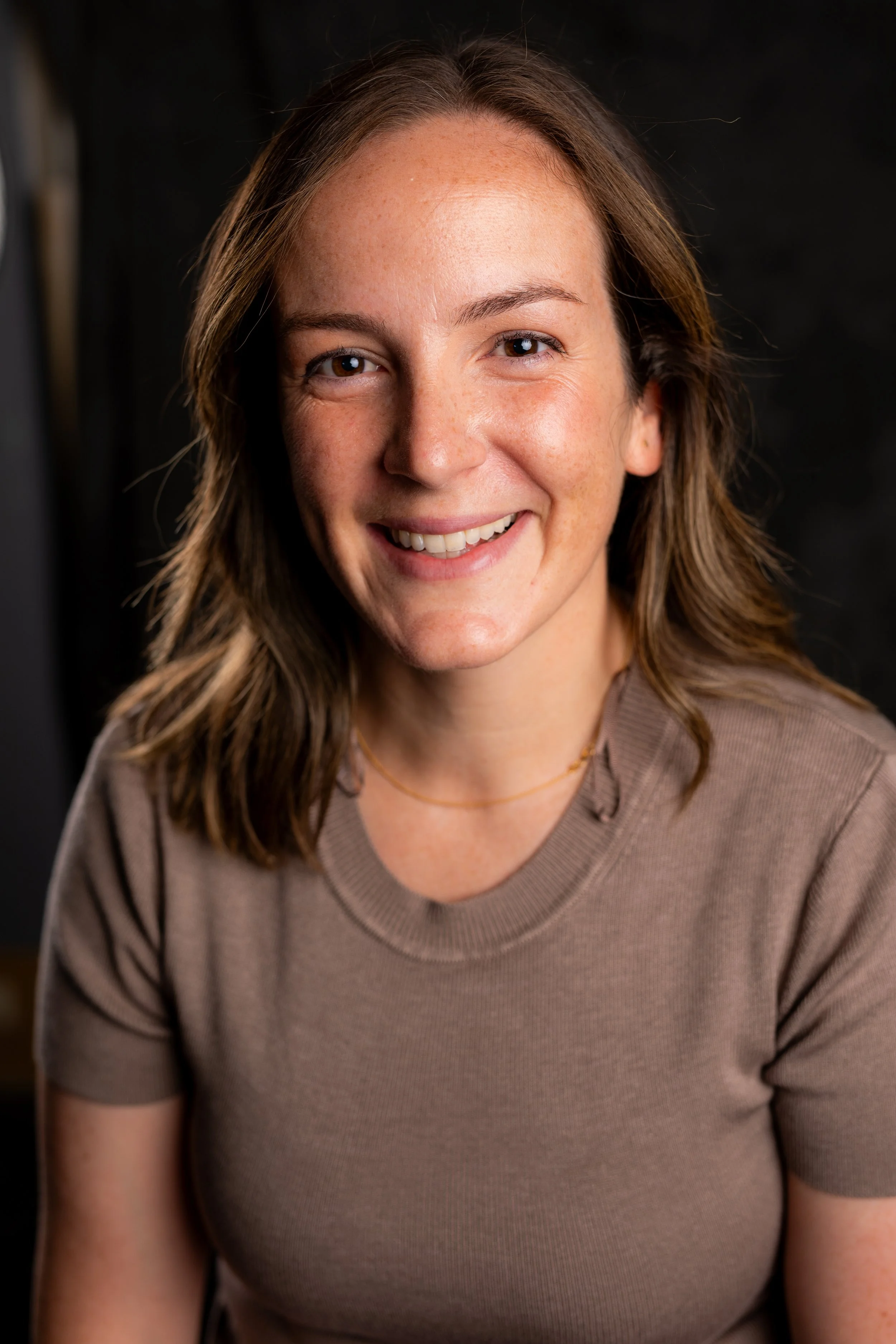 Close-up portrait of a smiling woman with shoulder-length brown hair, wearing a beige t-shirt, against a dark background.