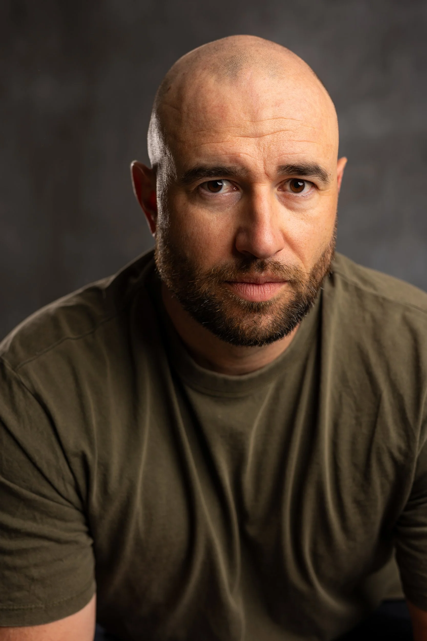 A close-up portrait of a bald man with a beard, wearing an olive-green T-shirt, looking directly at the camera against a dark, blurred background.