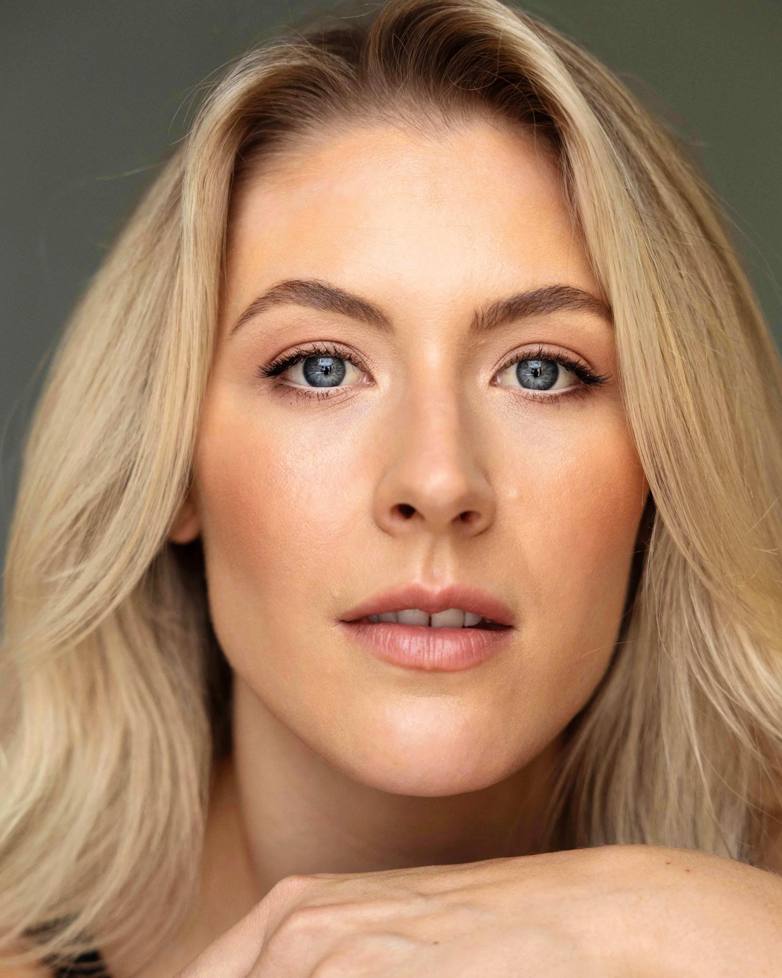 Close-up of a woman's face with blonde hair, blue eyes, and natural makeup, looking directly into the camera with a neutral expression.