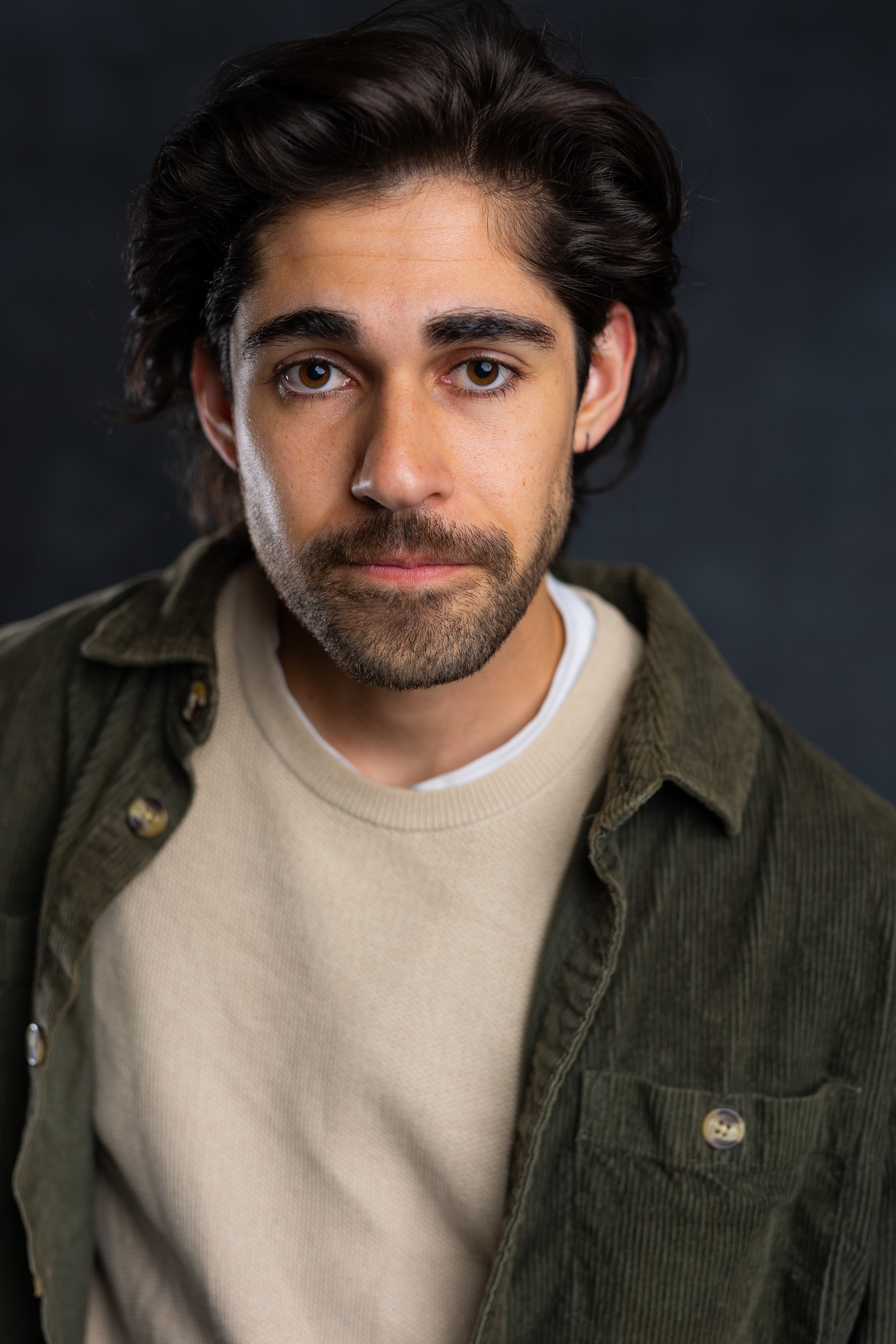 A young man with dark hair and a beard, looking into the camera, wearing a beige sweater and a green jacket, with a dark background.