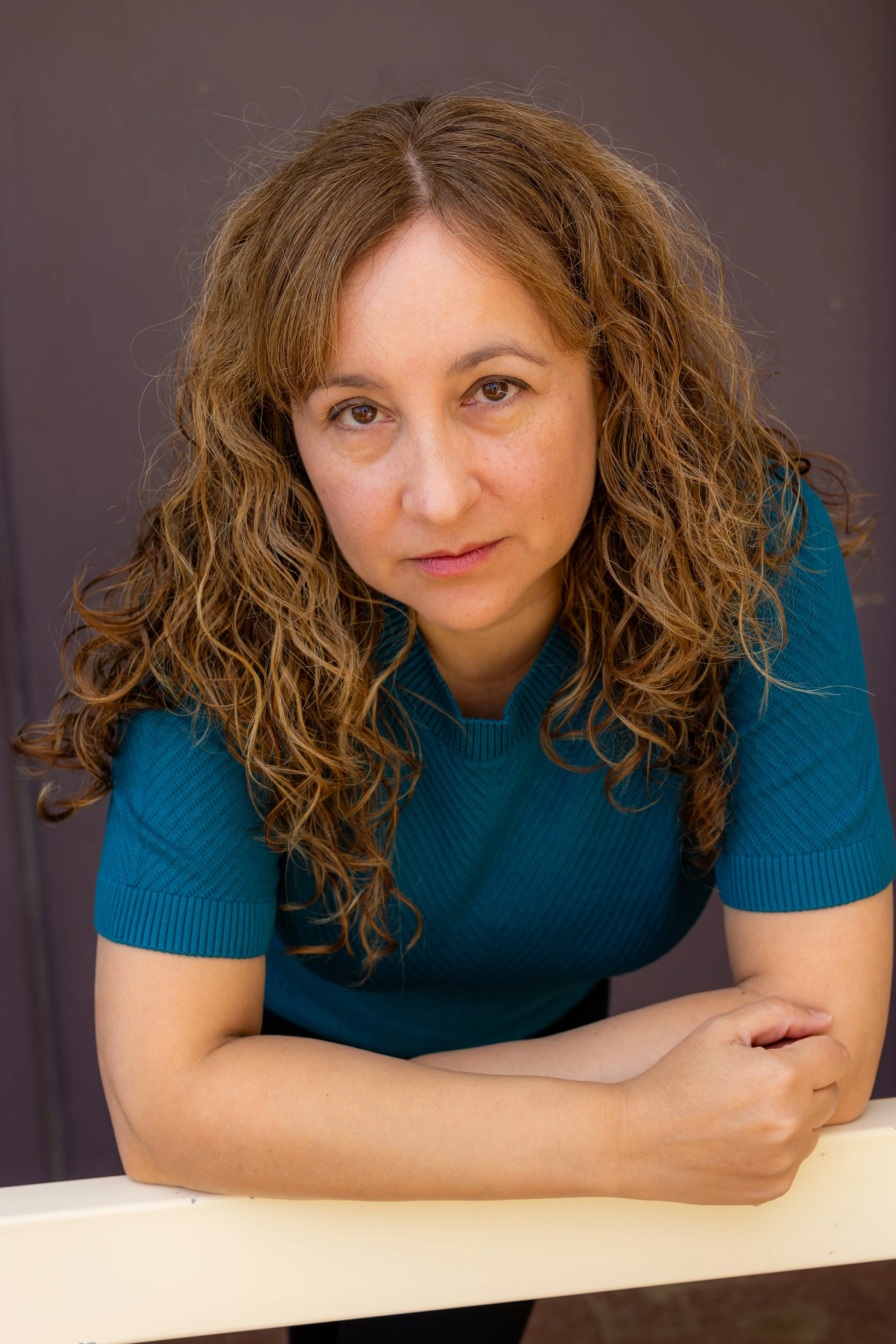 A woman with wavy, light brown hair and light skin wearing a teal short-sleeved top, leaning on a white surface and looking into the camera against a dark background.
