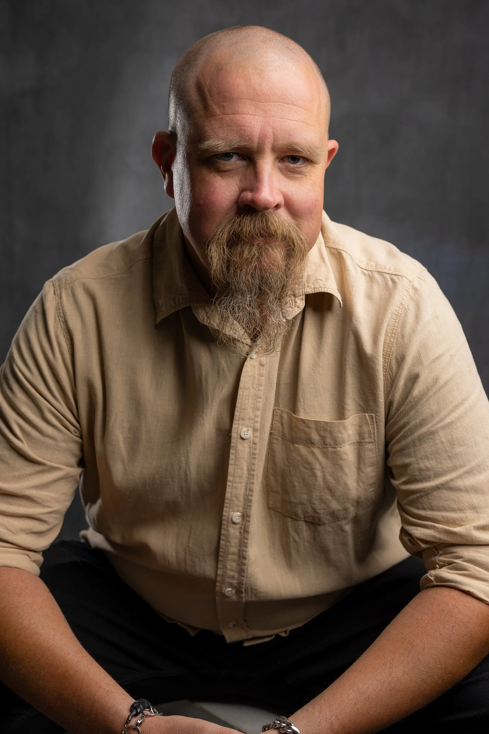 A man with a beard and shaved head wearing a beige shirt, sitting against a dark background.