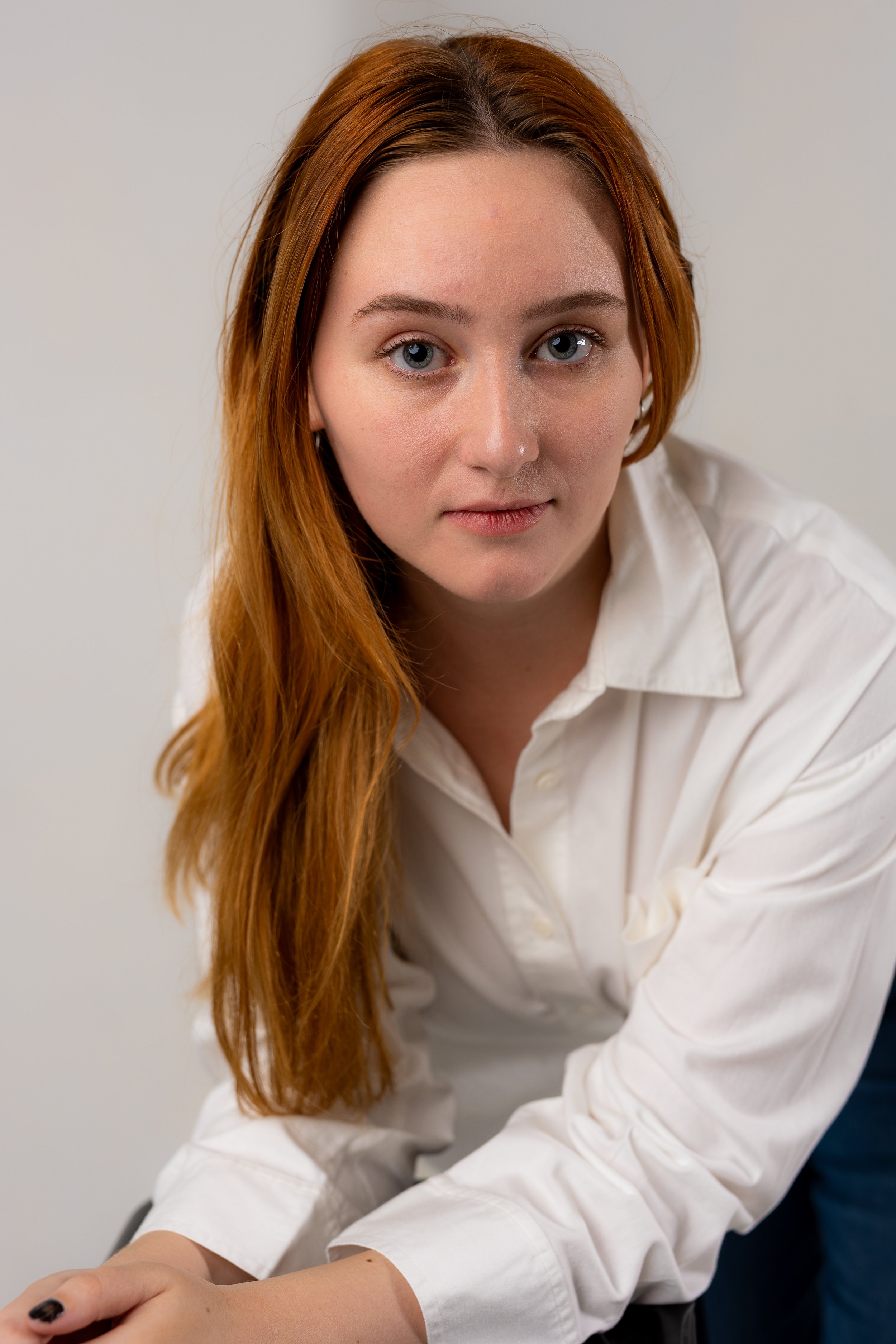 Close-up of a young woman with long red hair, wearing a white shirt, looking directly at the camera against a plain background.