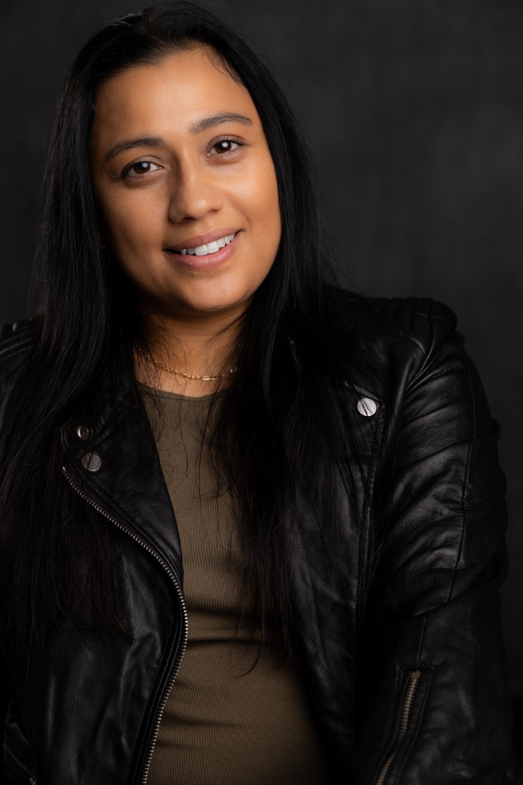 A woman with long black hair and light brown skin wearing a black leather jacket and a brown top, smiling at the camera against a dark background.