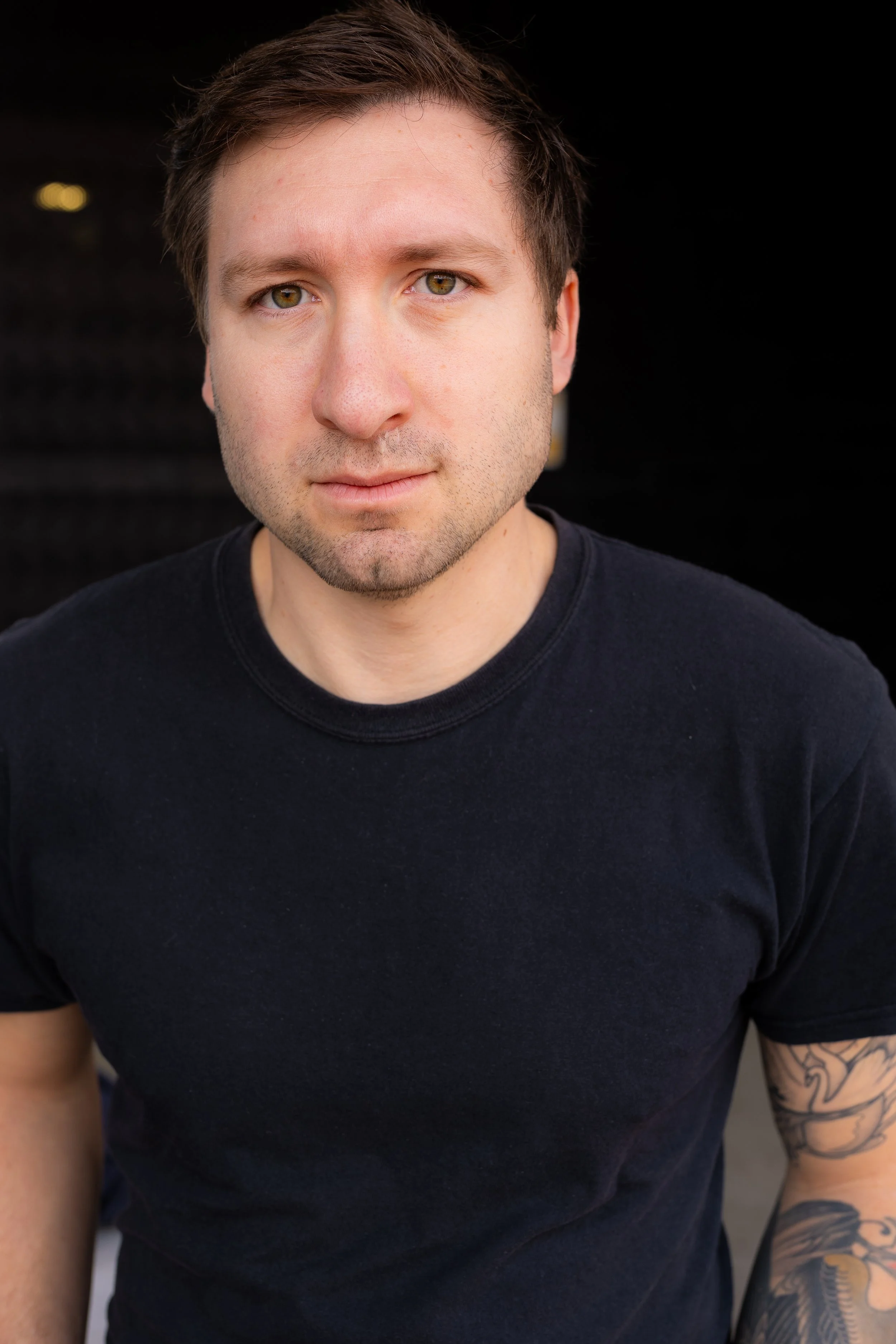 Close-up portrait of a young man with short brown hair, light skin, green eyes, and a slight stubble, wearing a black t-shirt, against a dark background.