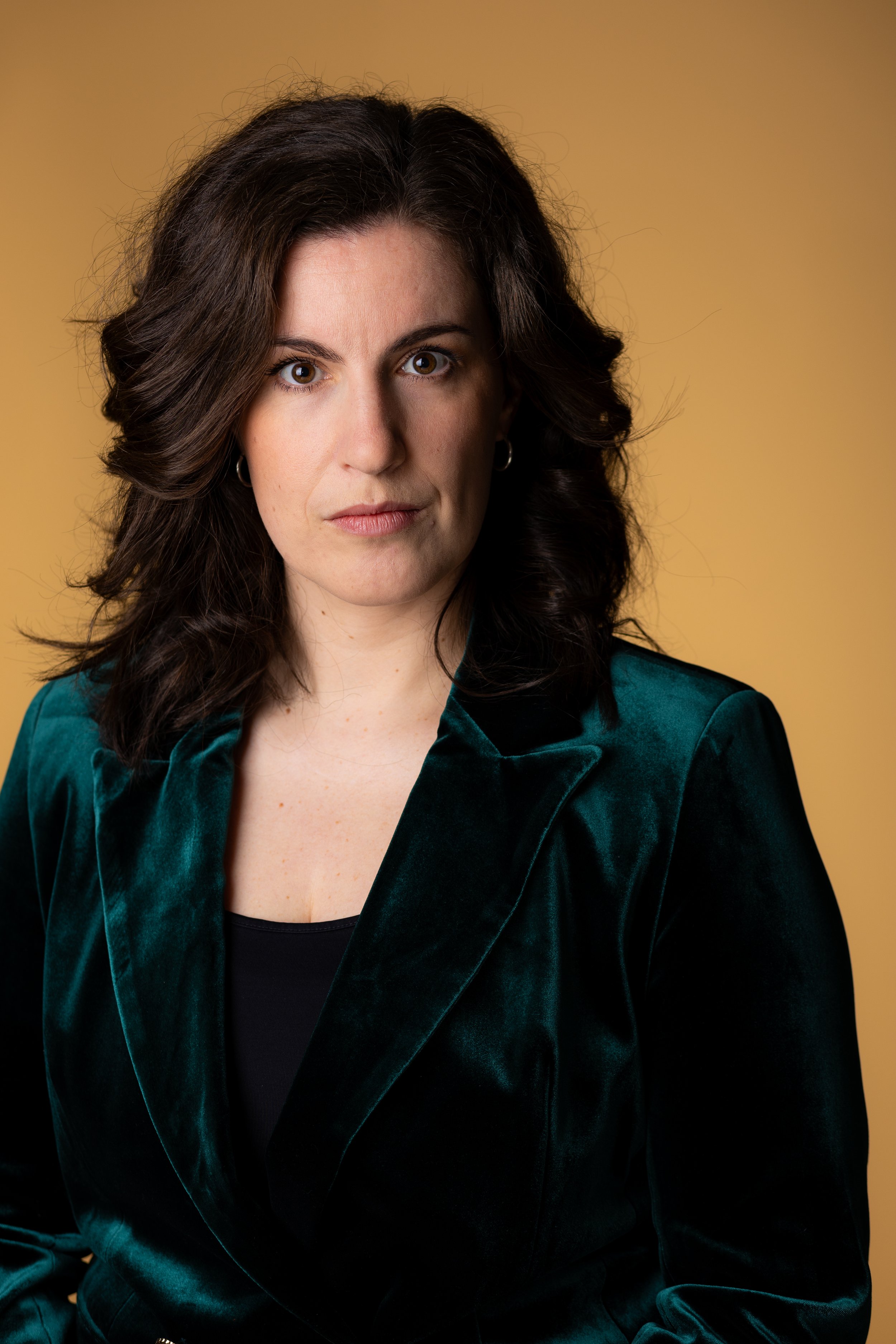 A woman with dark wavy hair wearing a dark green velvet blazer, looking directly at the camera against a yellow background.