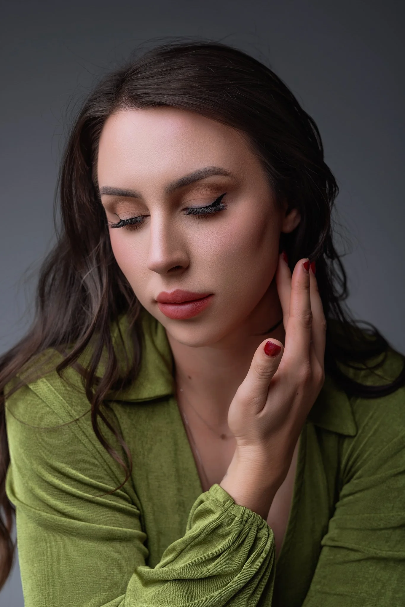 A woman with long brown hair, wearing a green top, with makeup and red nail polish, poses with her eyes closed and her hand touching her neck.