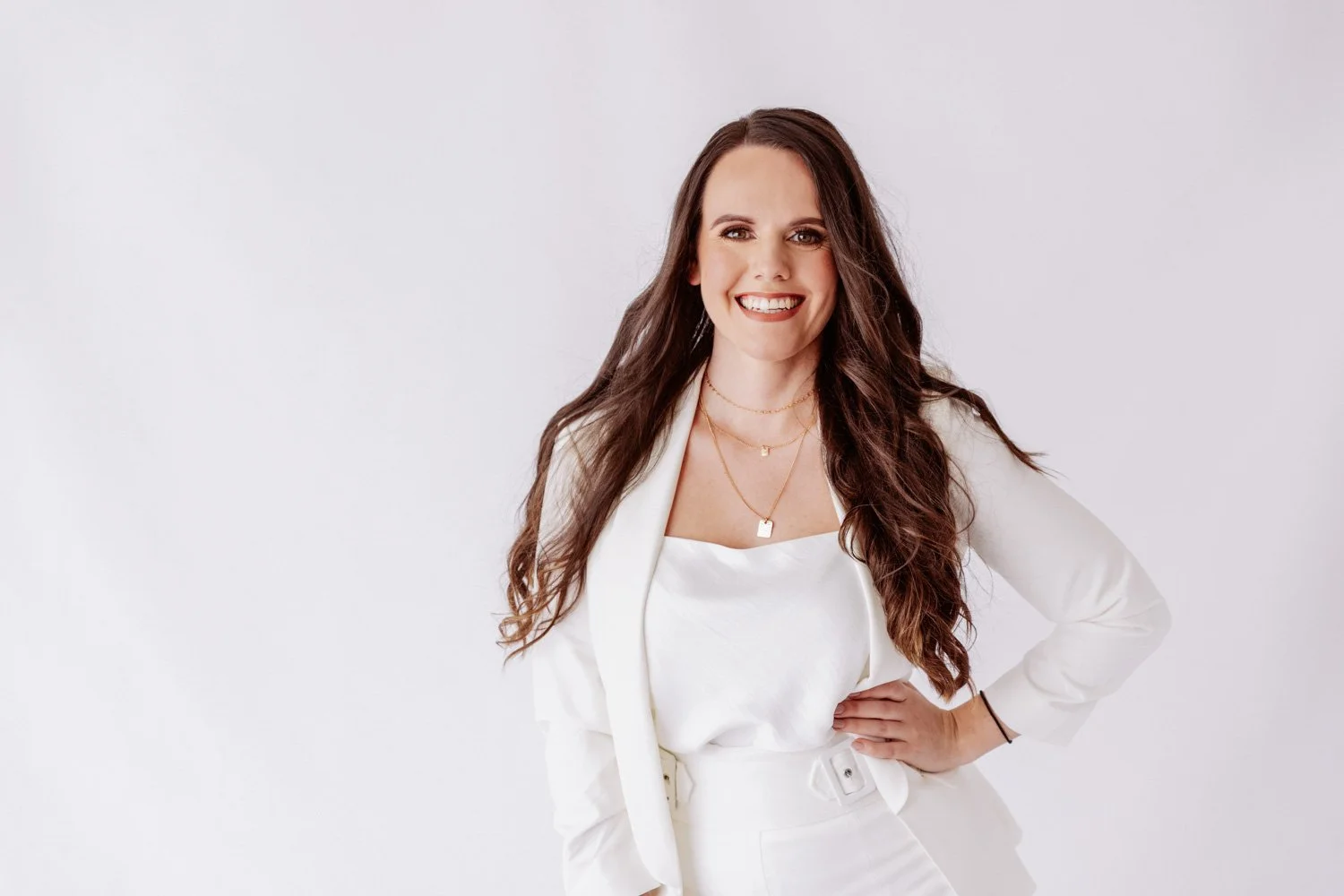A personal branding shoot of a woman smiling with long wavy brown hair, wearing a white blazer, white top, and layered gold necklaces, standing against a plain white background.