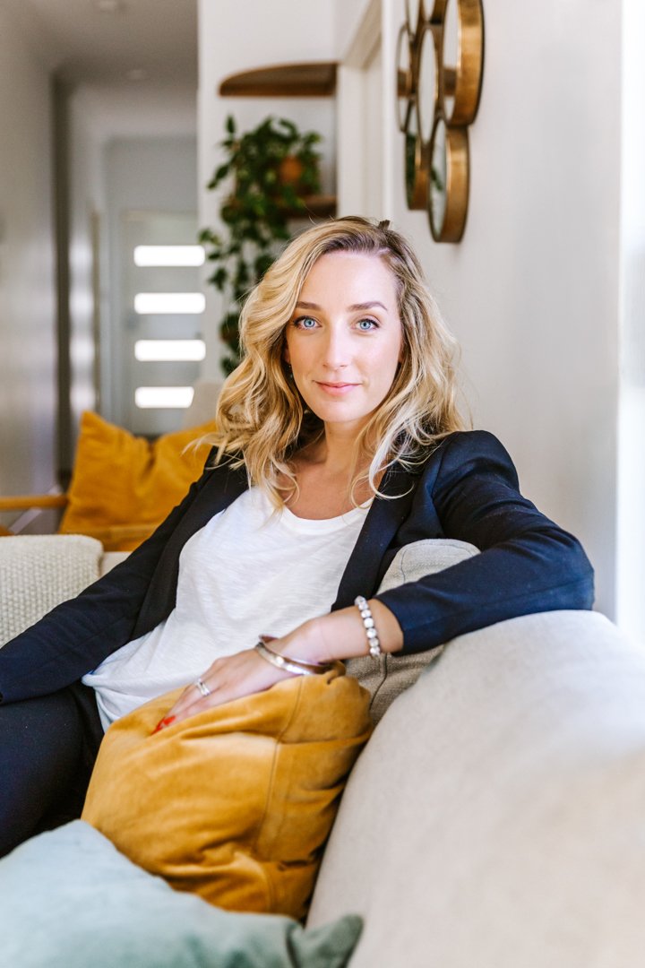 A woman with blonde curly hair, wearing a black blazer and white top, sitting on a beige sofa with colorful cushions in a living room.