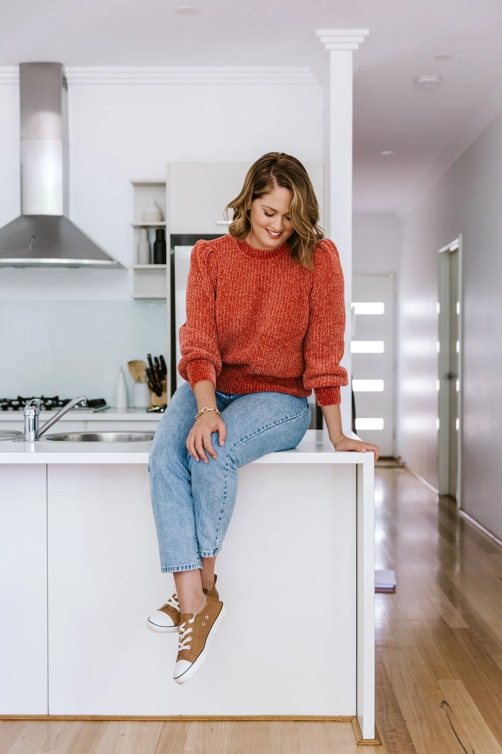 A smiling woman with shoulder-length brown hair wearing a red sweater, blue jeans, and tan sneakers, sitting on a kitchen counter in a bright, modern kitchen with white cabinets and wooden floors.
