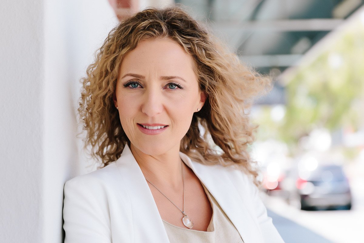 A personal branding shoot of a  woman with curly blonde hair, wearing a white blazer and a pendant necklace, outdoors.