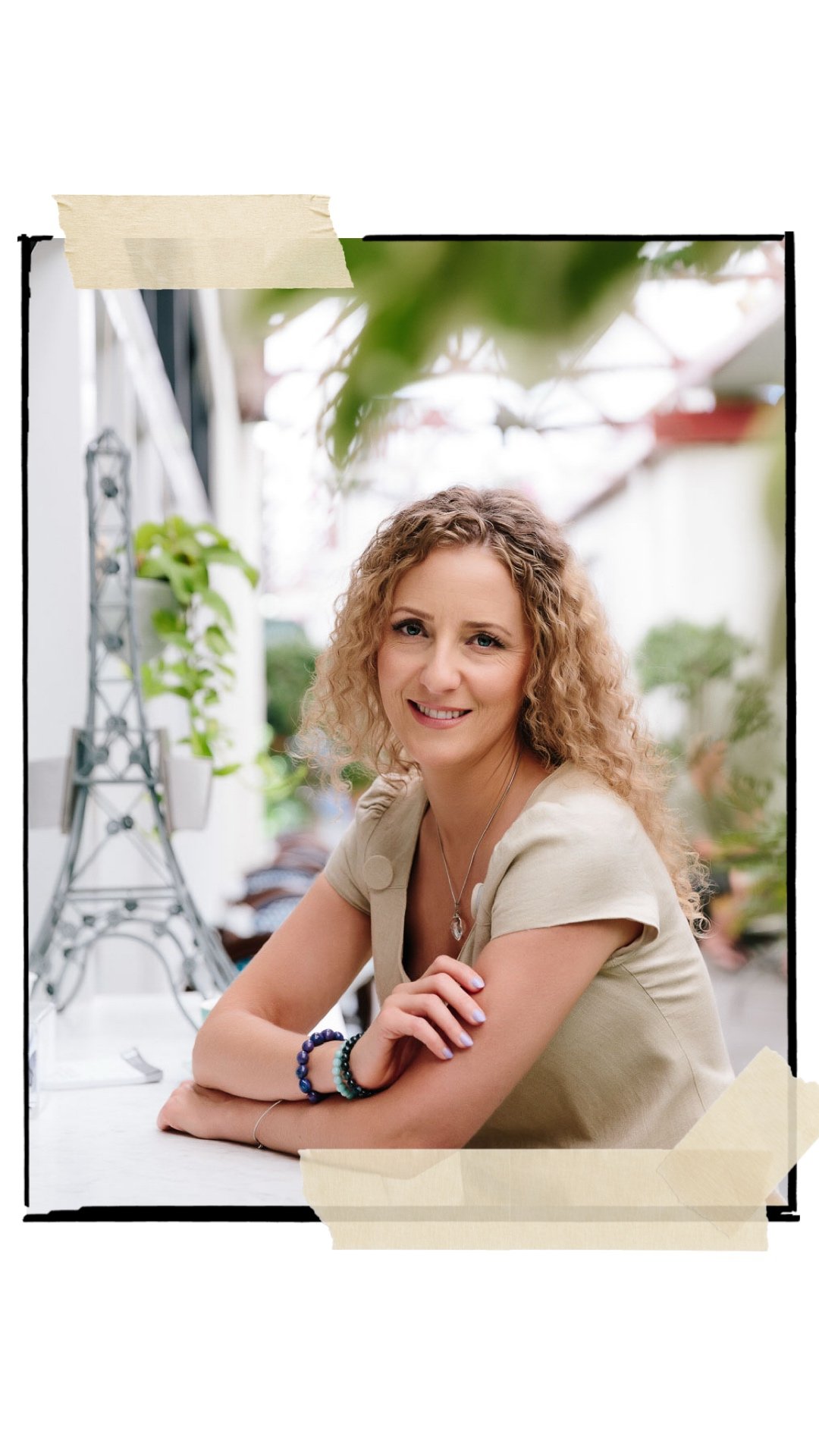 A woman with curly blonde hair sitting at a table outdoors, smiling at the camera, with greenery in the background.