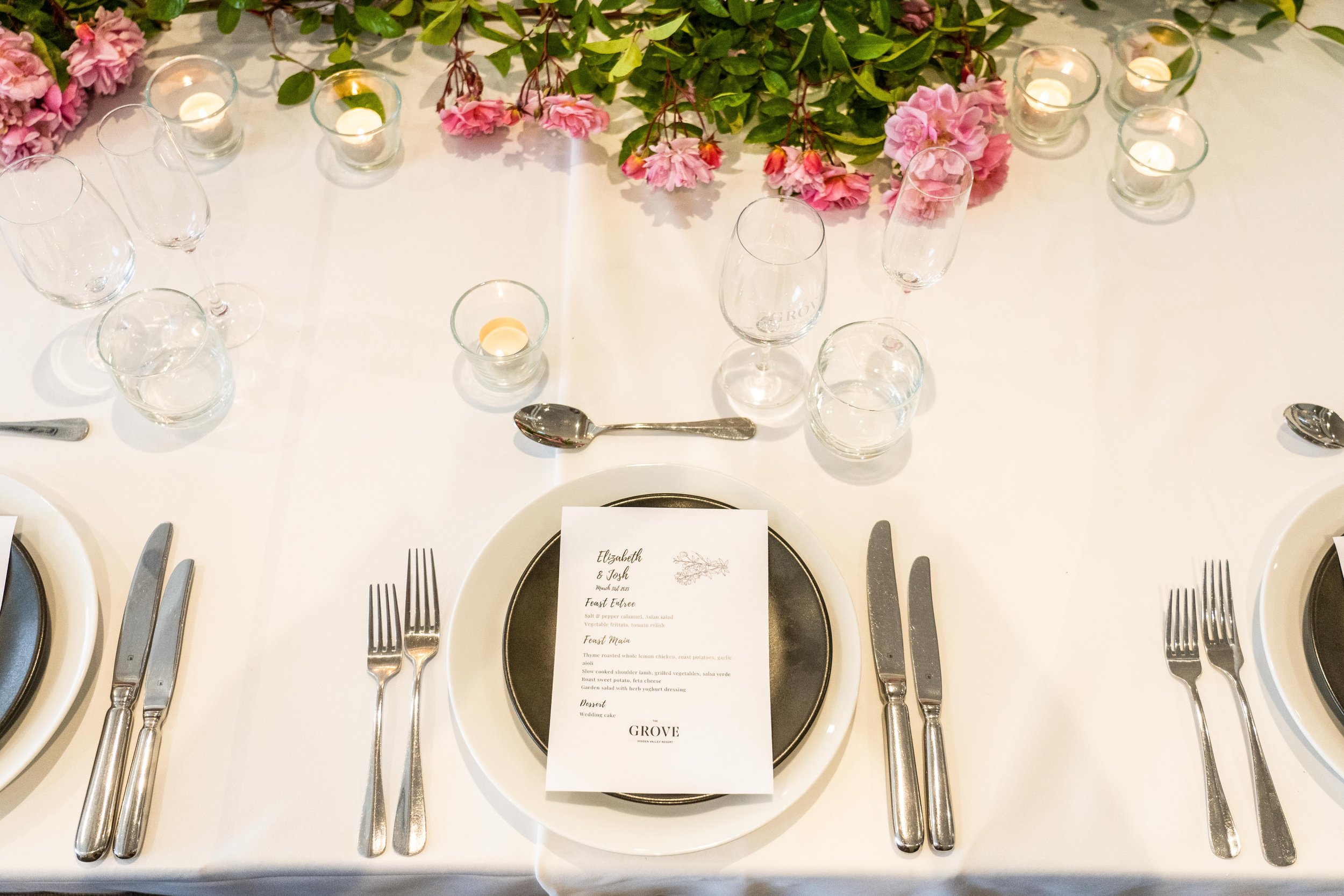 Elegant table setting with white tablecloth, multiple wine and water glasses, silverware, a menu card, pink flowers, green leaves, and small tea light candles.