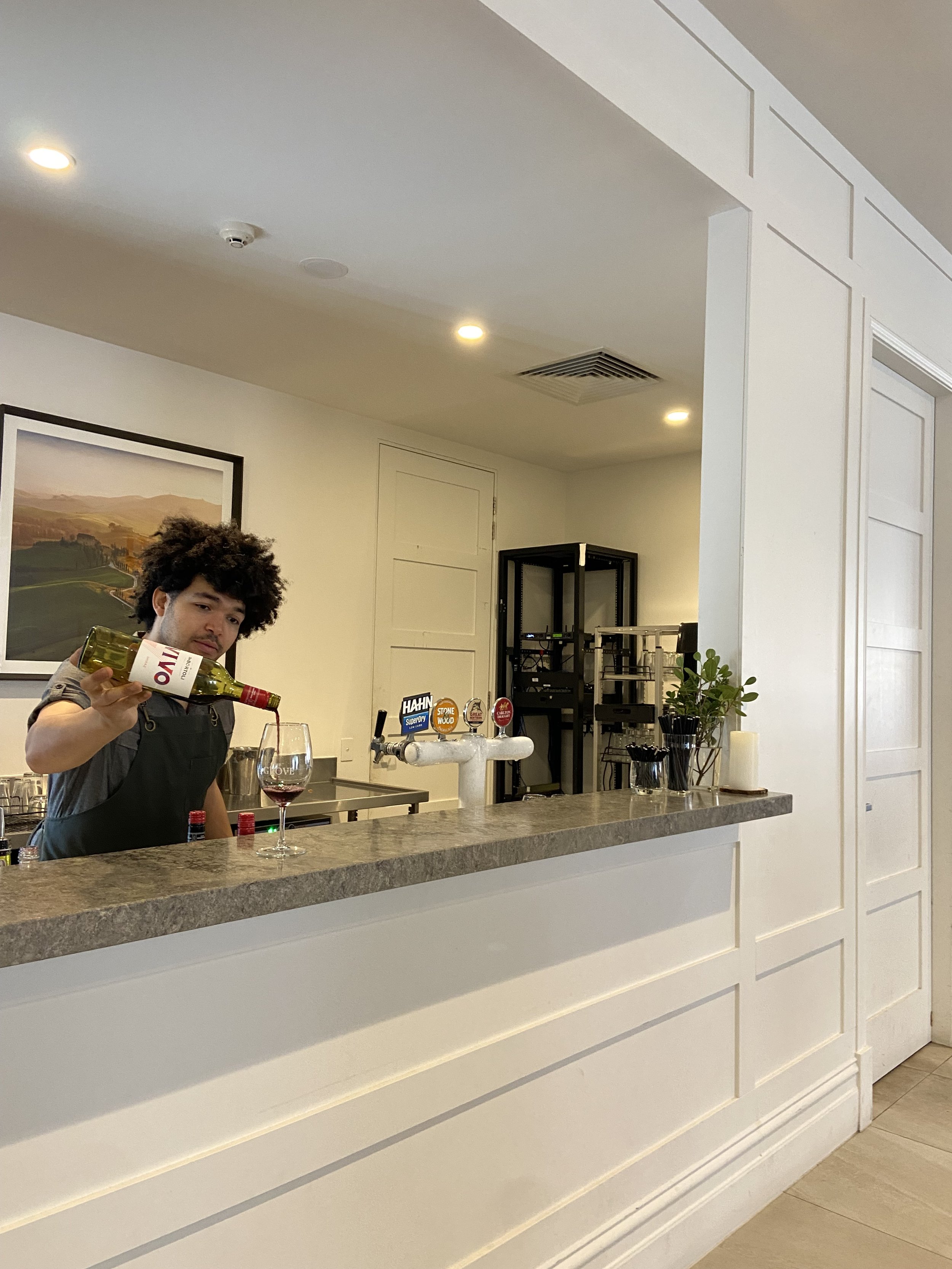 A bartender pours red wine into a glass behind a bar counter in a well-lit room.