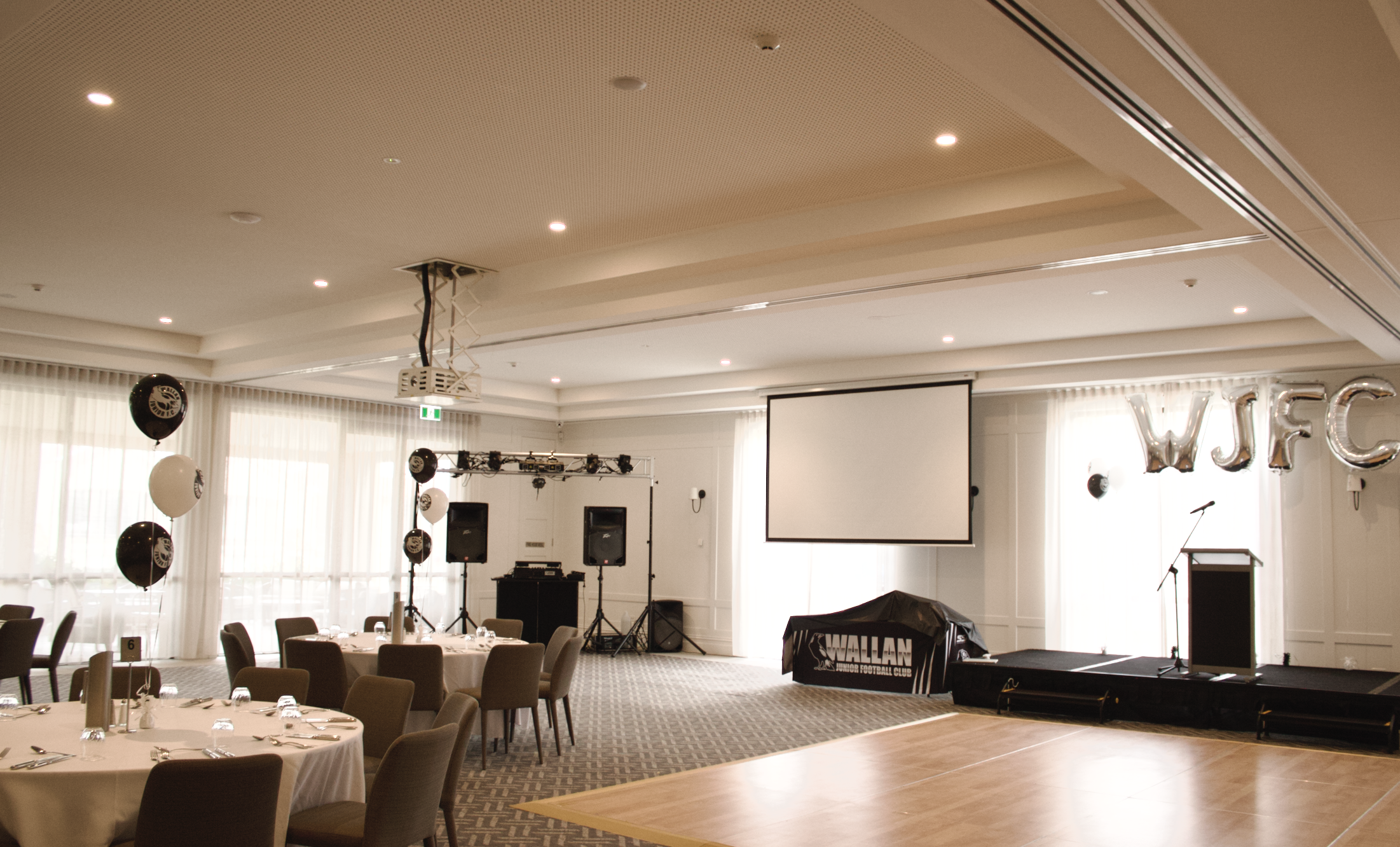 Indoor event space with round tables, chairs, a stage with a podium, large projection screen, sound equipment, and silver balloons spelling 'WJFC'