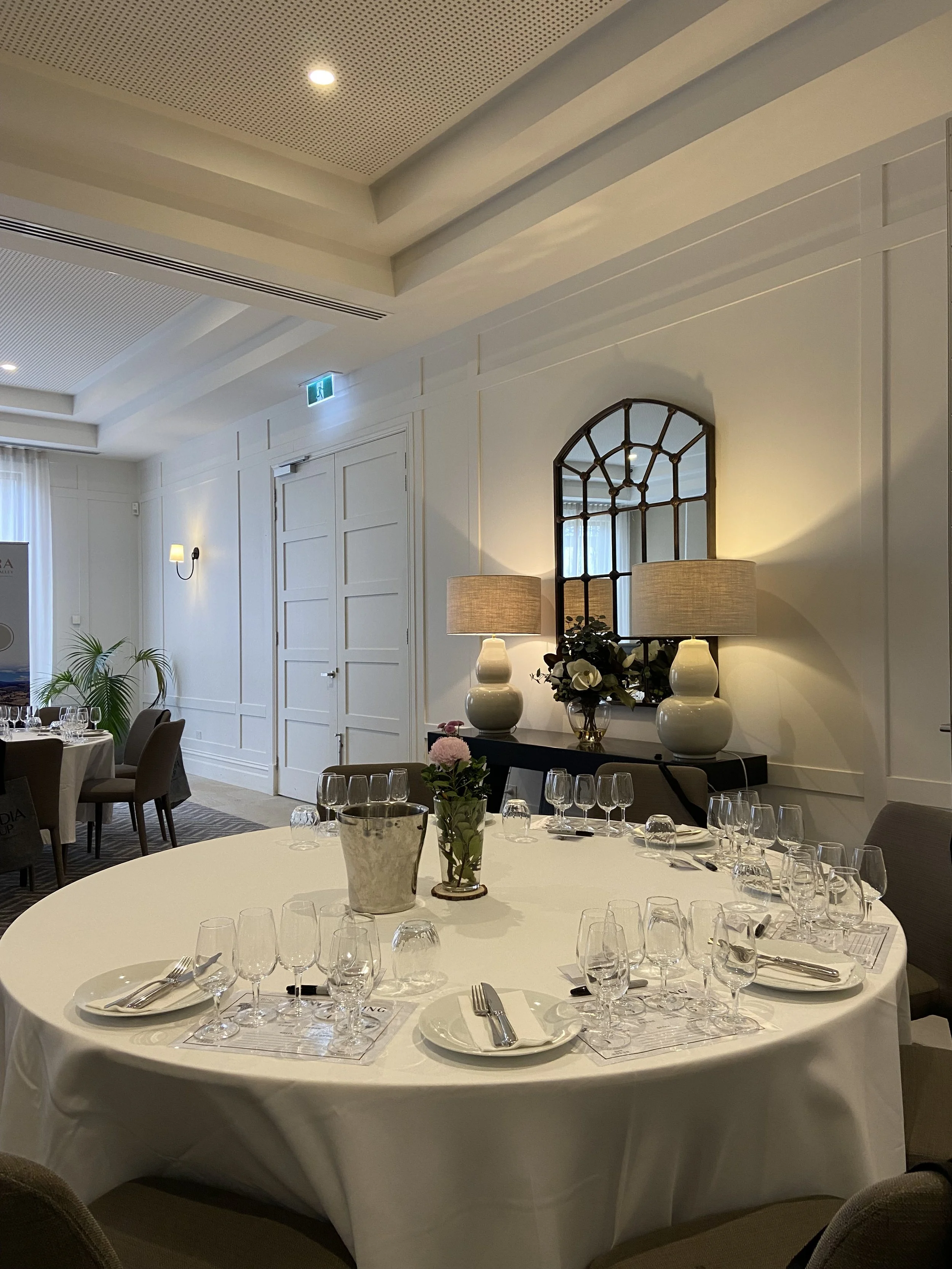 Round banquet table set with wine glasses, water glasses, cutlery, and napkins, in an elegantly decorated room with white paneled walls, lamps, floral arrangements, and a large mirror.