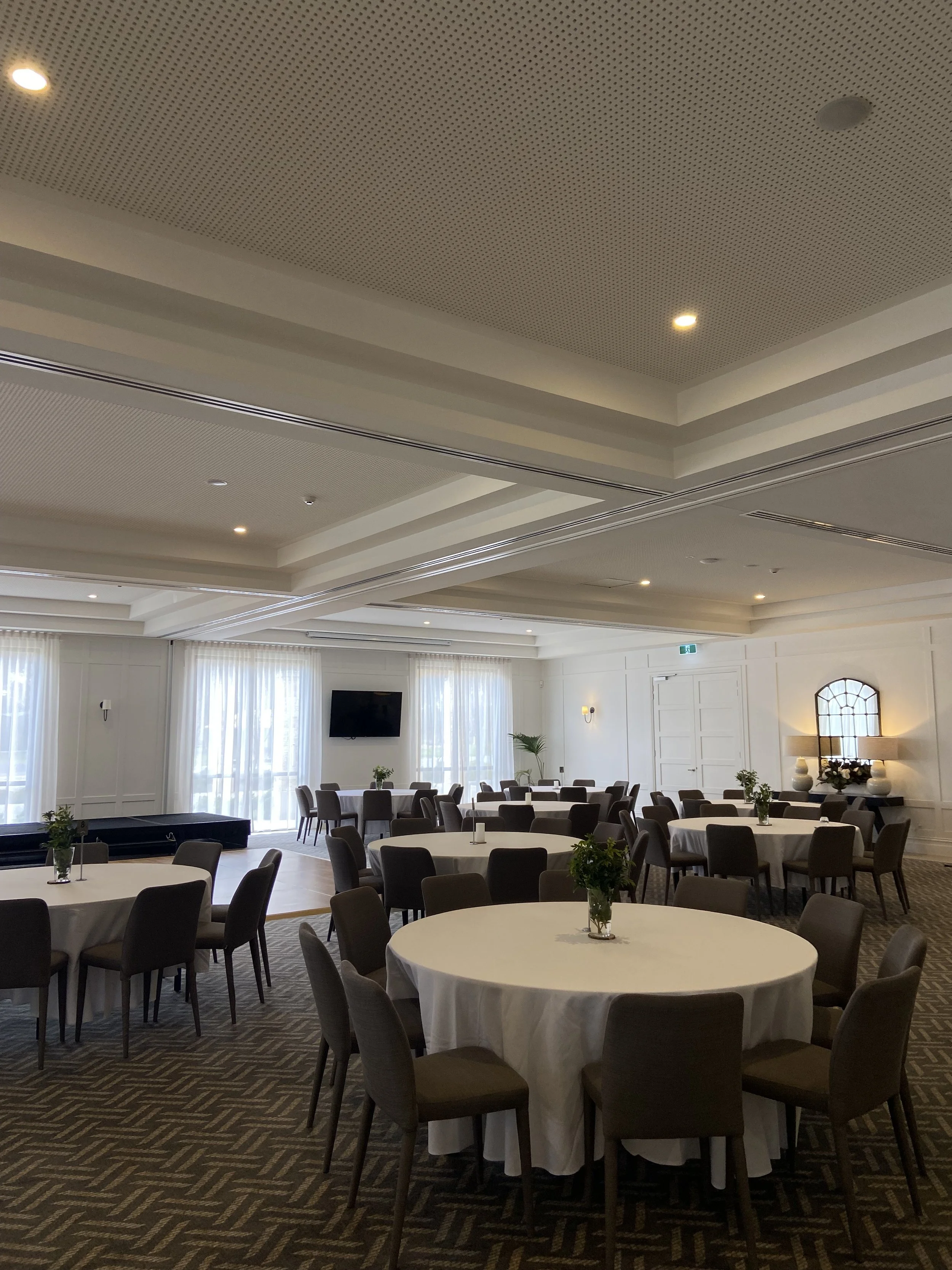 Elegant event space with round tables covered in white cloth, brown chairs, and small green plant centerpieces. Soft natural light filters through sheer curtains, and a flat-screen TV is mounted on the wall. Decor includes vases and lamps, and the ceiling has recessed lighting.