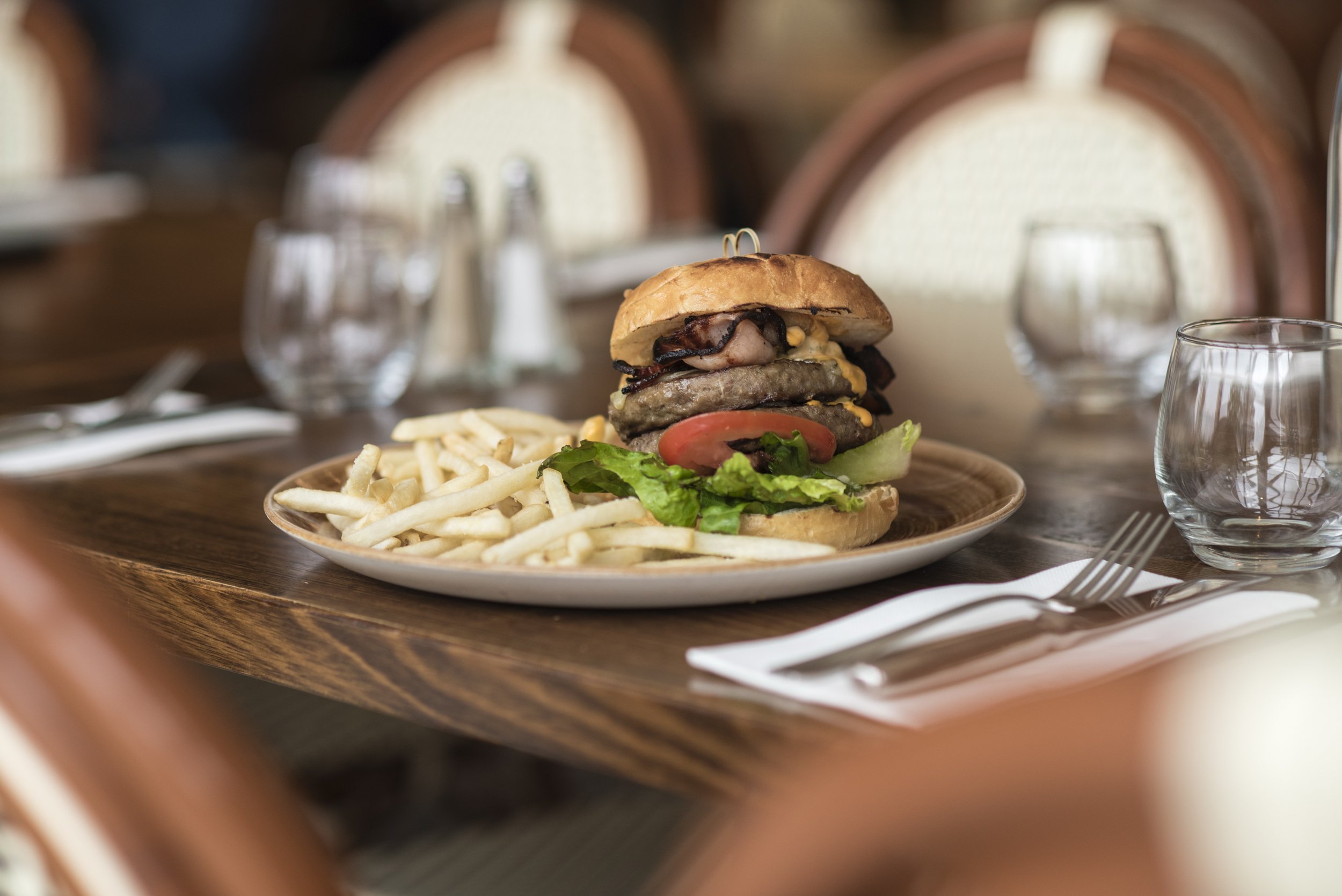 A plate of French fries and a large burger with lettuce, tomato, cheese, two beef patties, bacon, and a bun, on a wooden table with empty glasses and utensils in a restaurant setting.