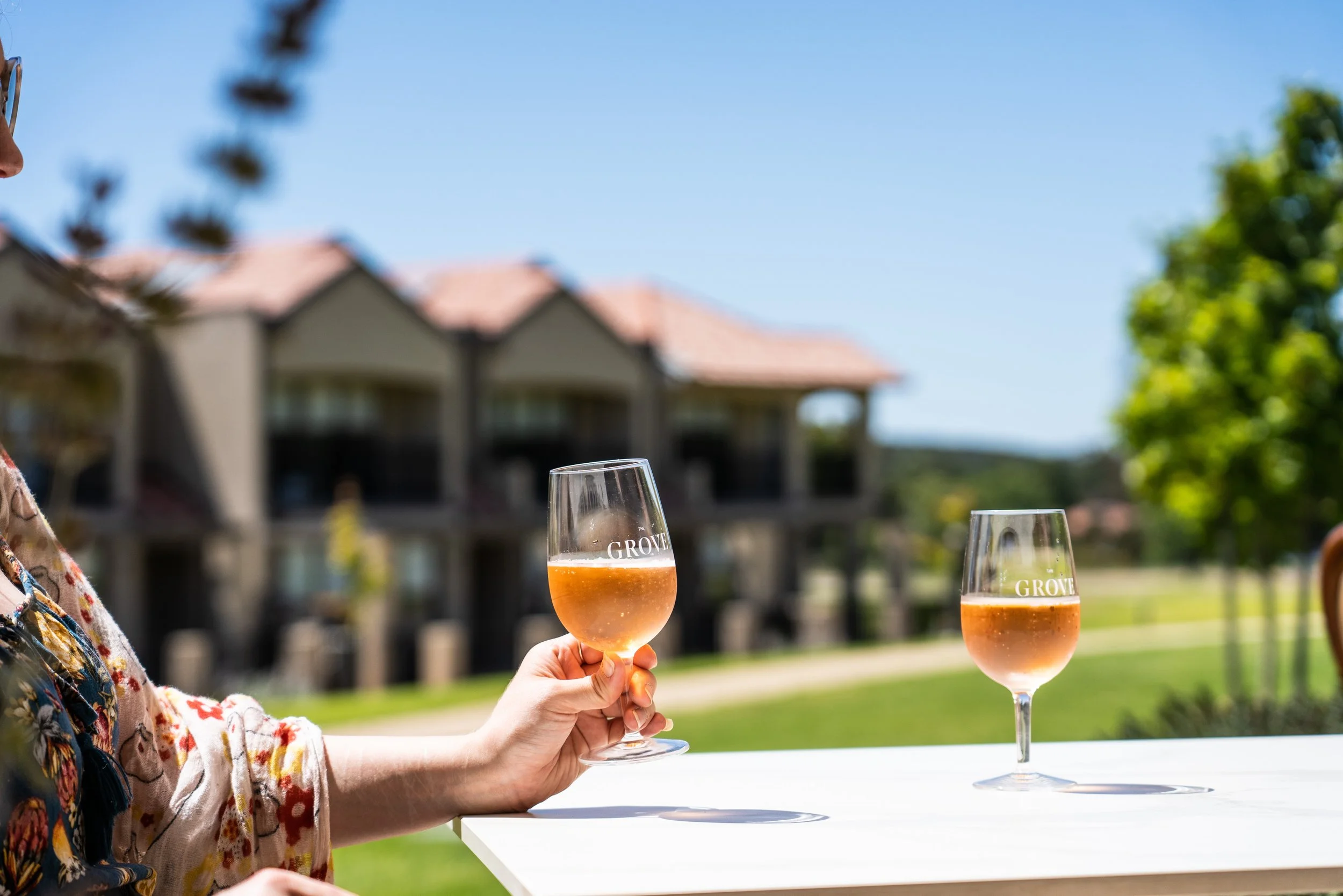 Person holding a wine glass with pink wine on a white table outside, with two more glasses and residential buildings in the background under a clear blue sky.