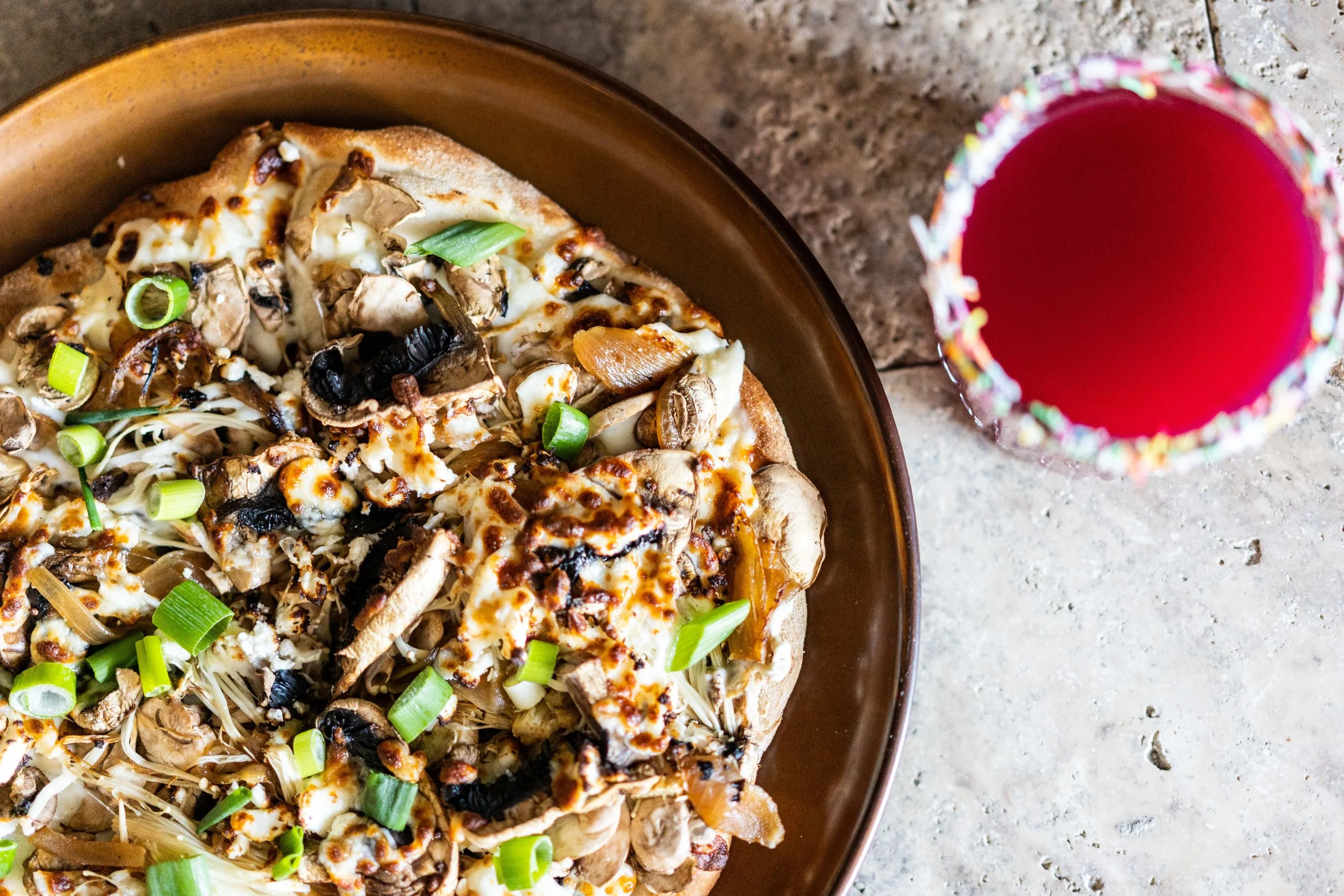 Pizza topped with mushrooms, green onions, and cheese next to a glass of red beverage with a colorful rim.