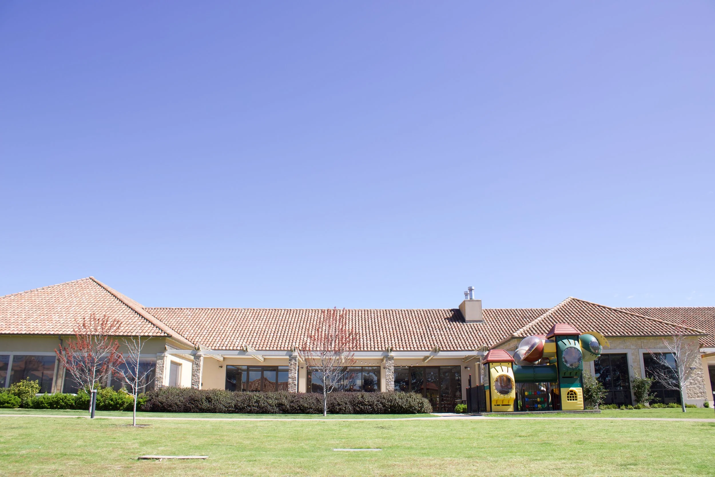 Single-story building with a red-tile roof, large windows, and a playground structure outside, with a green lawn and a clear blue sky.