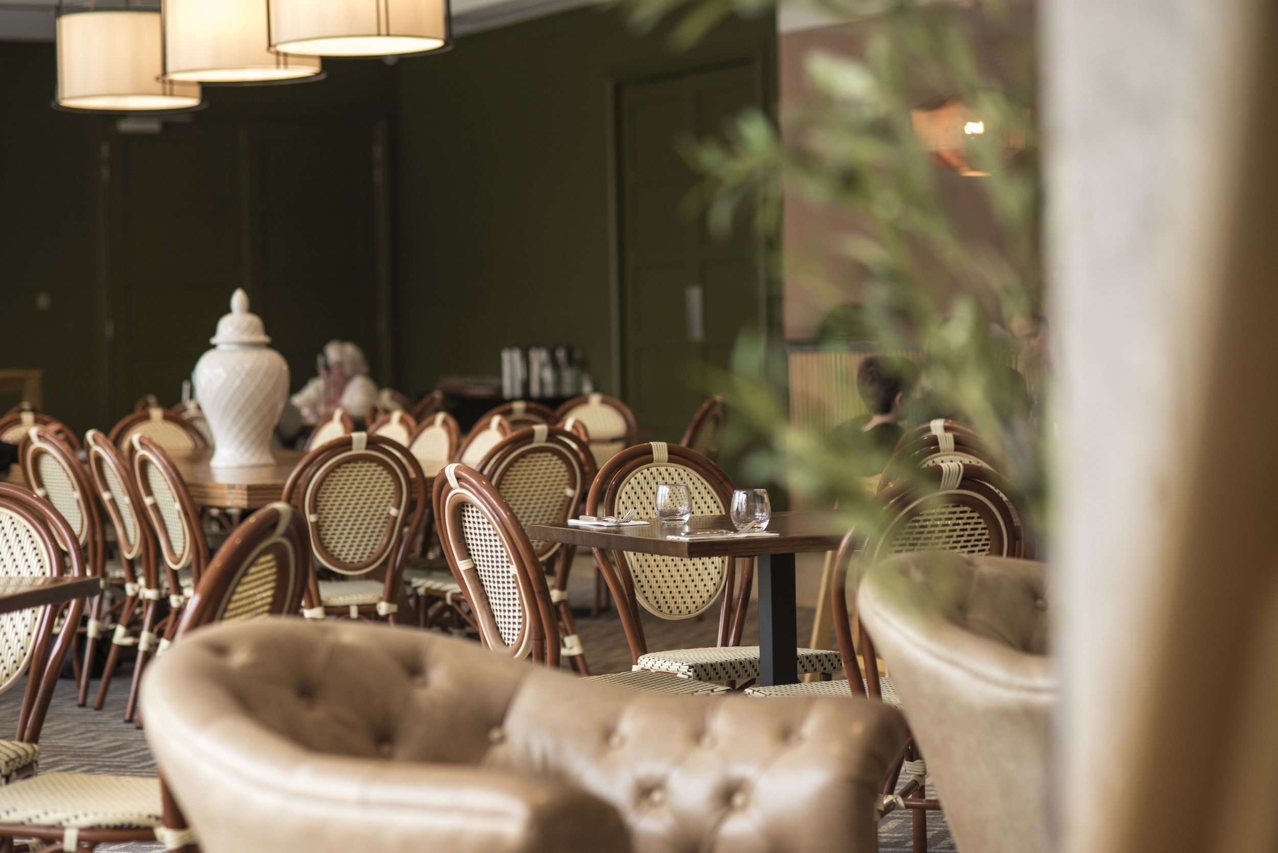 Empty restaurant dining area with wooden tables, beige woven chairs, and a large white decorative vase, with a green wall and window in the background.